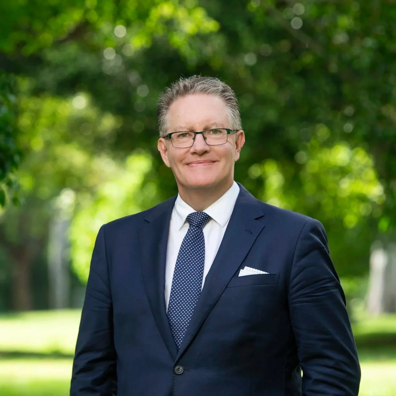 A man in a dark suit, white shirt, and patterned tie standing outdoors with trees and greenery in the background.