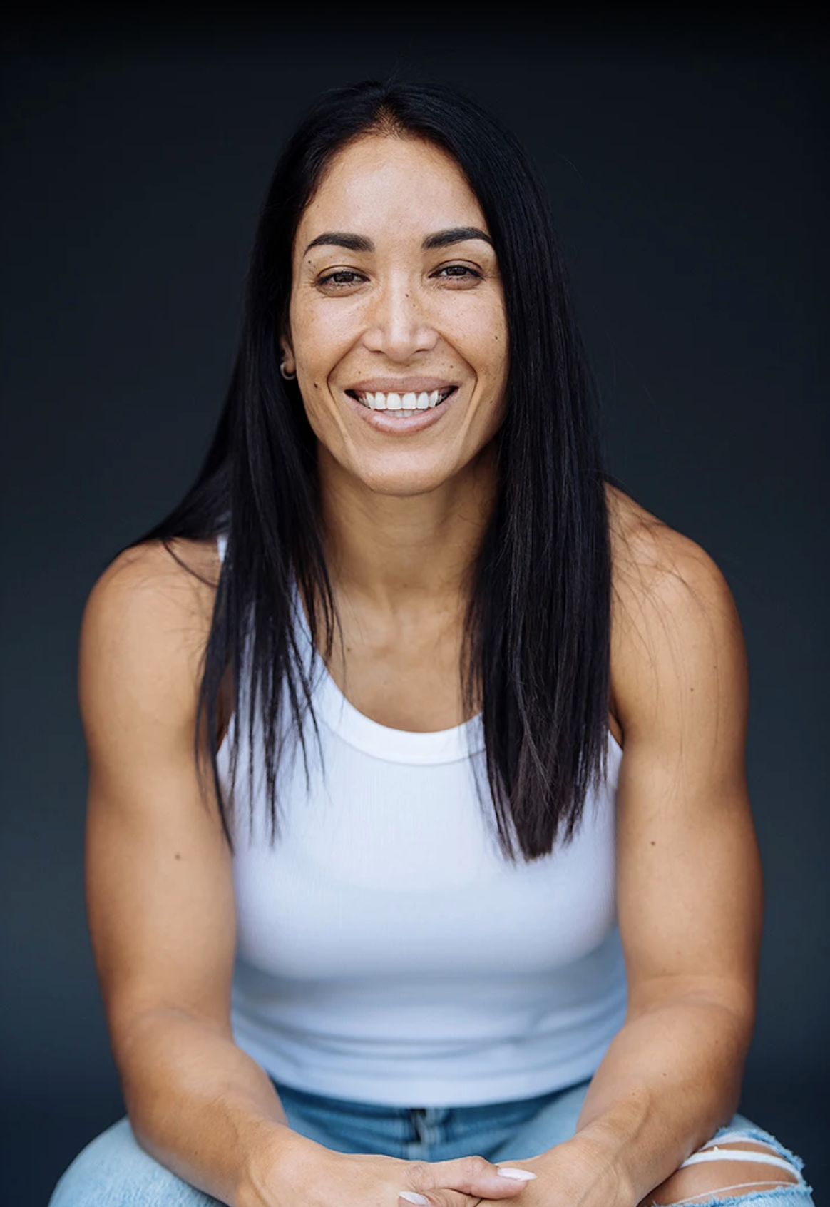 Portrait of a woman with long black hair, smiling, wearing a white tank top and ripped jeans, against a dark background.