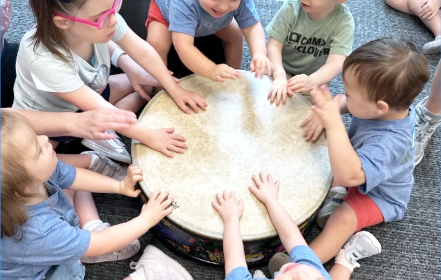 Children gathered around a round drum, reaching out to touch its surface.