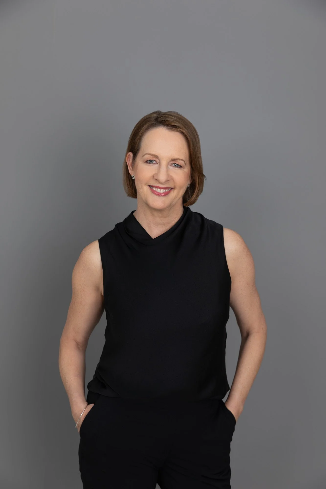 A woman with shoulder-length reddish-brown hair, wearing a sleeveless black top, smiling, standing against a gray background.