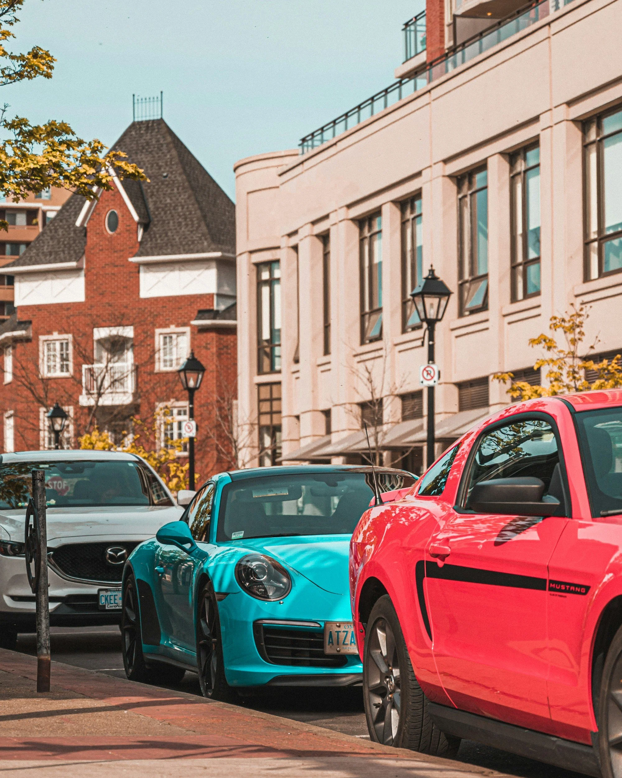 A street scene with parked cars, including a blue sports car, a red Mustang, and a white Mazda, lined up along a sidewalk with buildings and trees in the background.