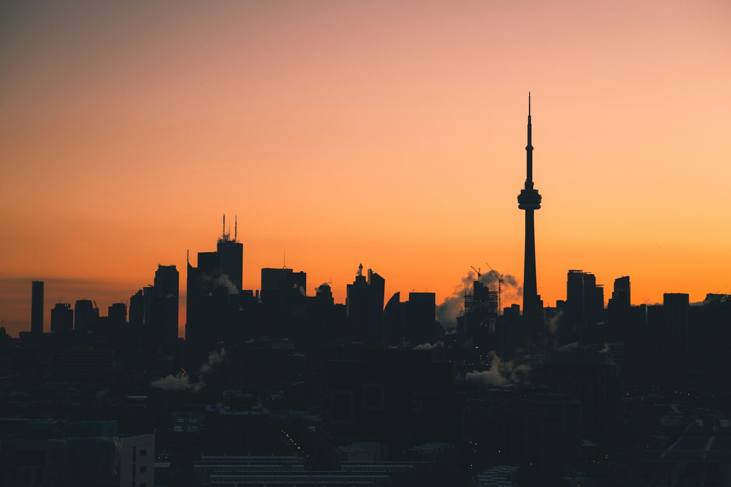 Silhouette of a city skyline with a tall television tower at sunset, with orange and pink sky in the background.