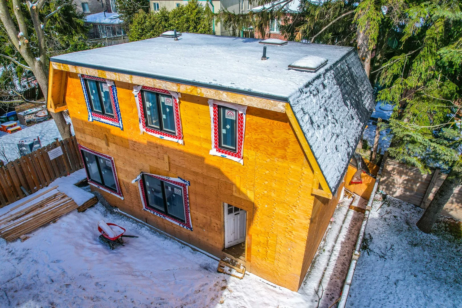 Wooden house under construction with visible framing and plywood walls, surrounded by trees and a blue sky.