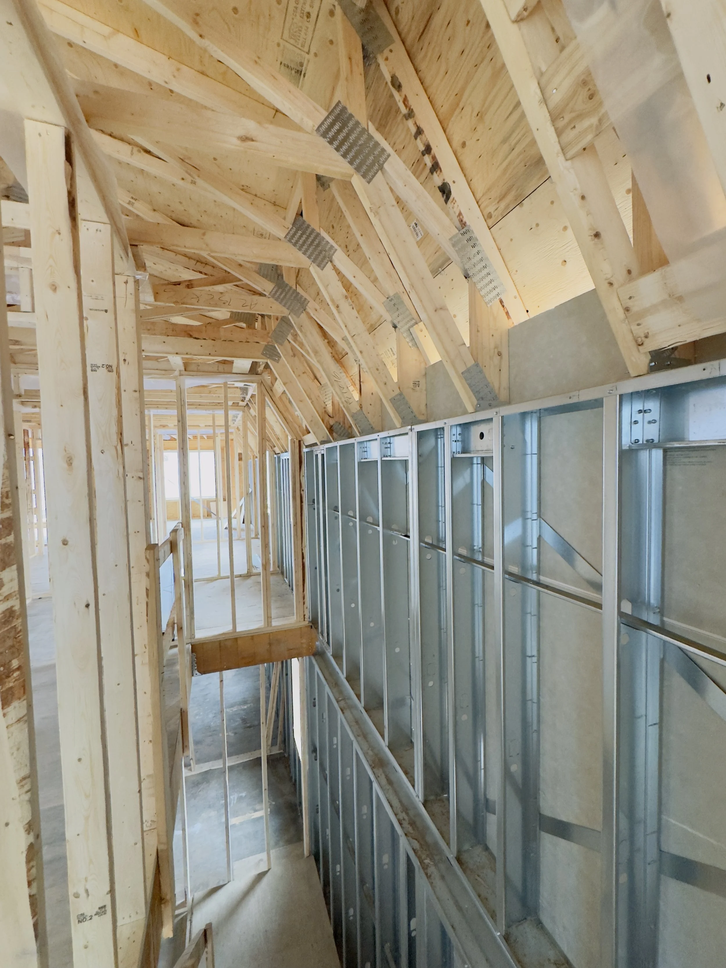Construction site where a steel framework is being assembled between a brick building and a yellow wall, with a ladder and various tools and supplies scattered on the ground, under a clear blue sky.