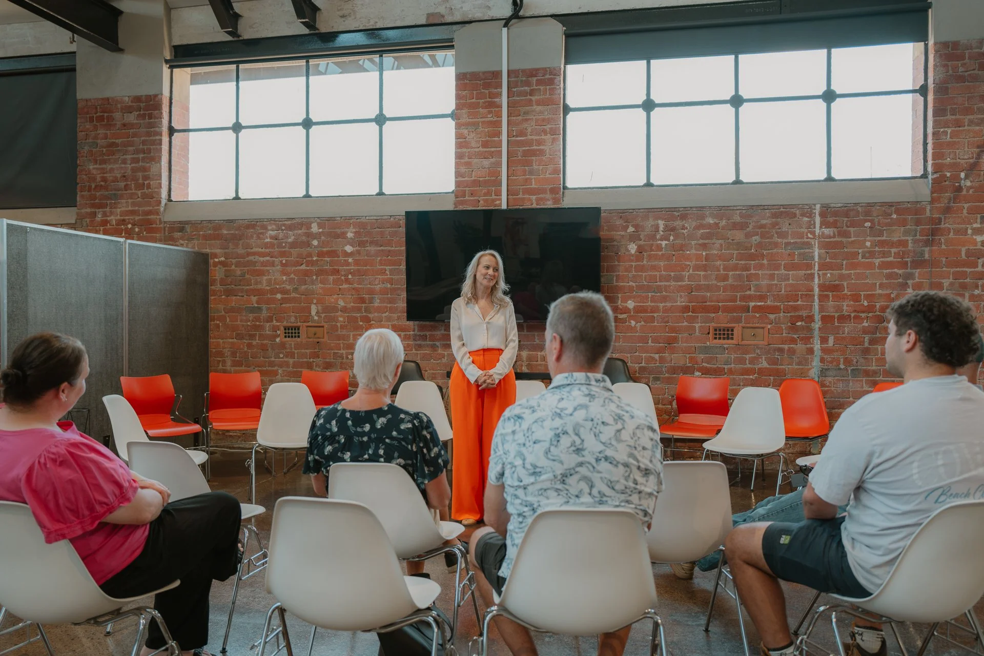 A woman giving a speech or presentation to a small audience in an industrial-style room with brick walls and large windows.
