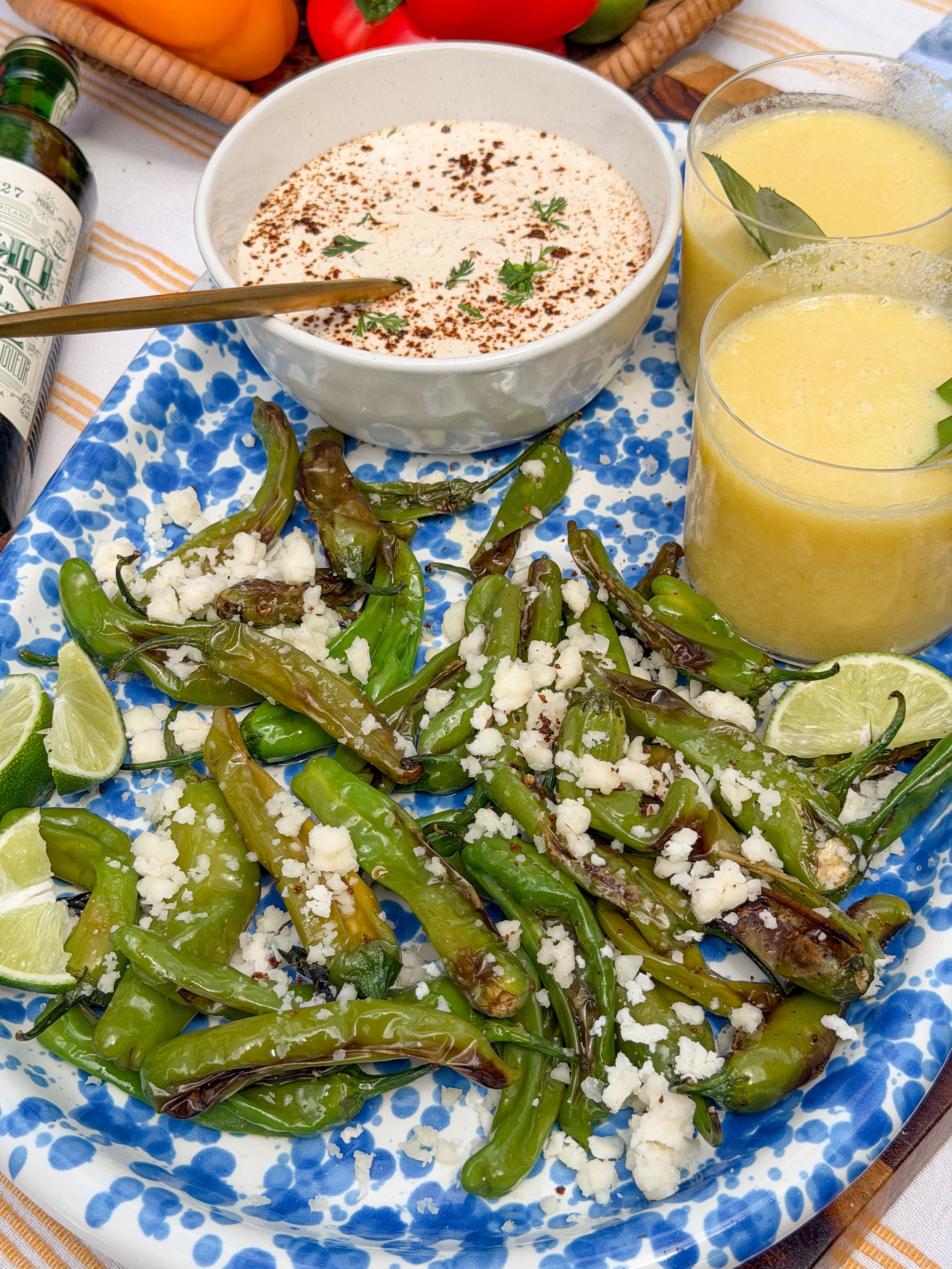 small green peppers sit on a blue speckled platter next to two cocktails with a pineapple margarita. The peppers are sprinkled with cotija cheese and sitting next to a bowl of elote sauce sprinkled with red pepper.