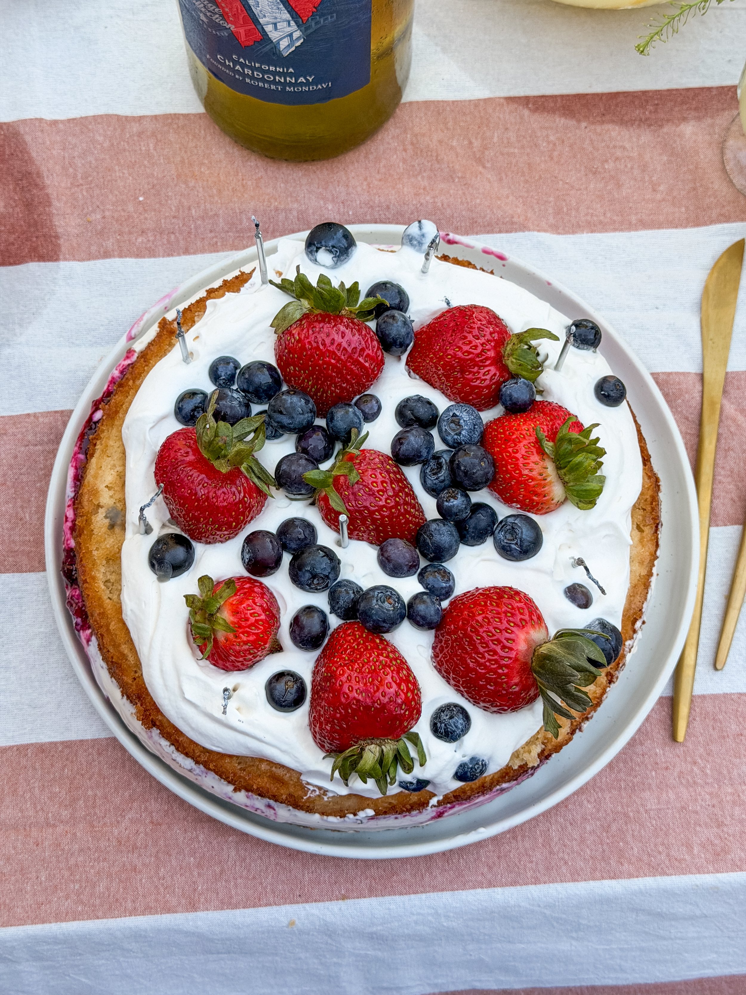 Round cake with strawberries and blueberries sitting on a bed of whipped cream. Cake is on a red and white striped tablecloth.