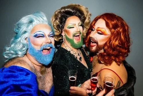 Three people dressed in colorful drag costumes sharing a joyful moment, each holding a glass of what appears to be champagne or sparkling wine, smiling and laughing.
