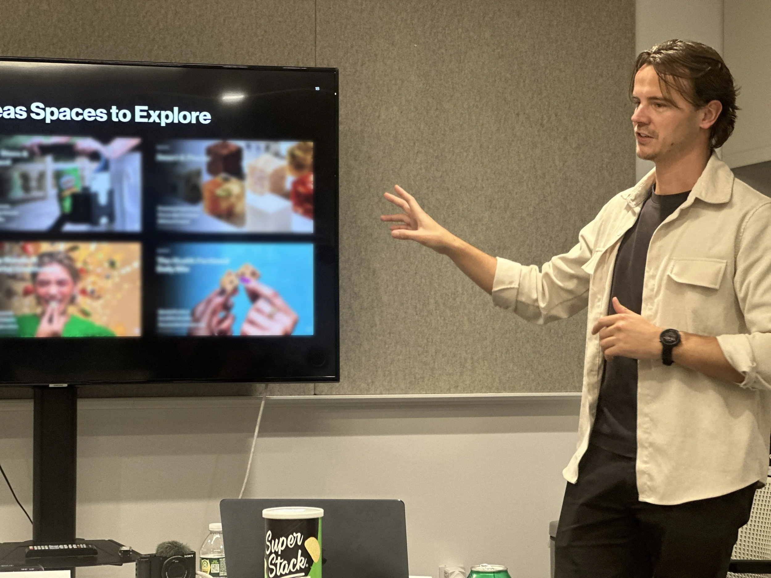 A man with brown hair and a beige jacket giving a presentation in front of a large screen displaying colorful images and text. A table with a black Super Stack coffee cup, a water bottle, and other items is in the foreground.