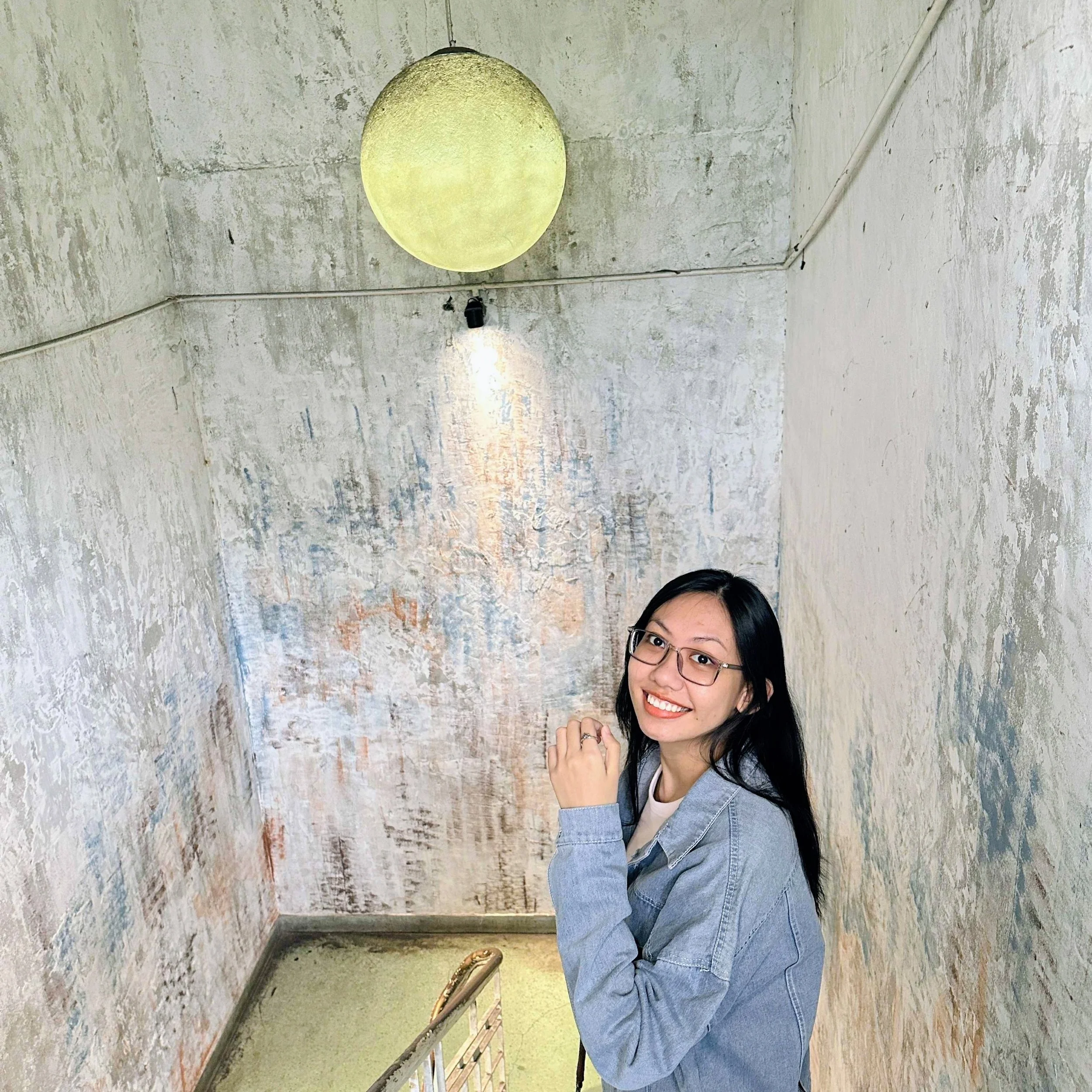 A young woman with glasses and a denim jacket smiling in a small, distressed textured room with a round ceiling light.