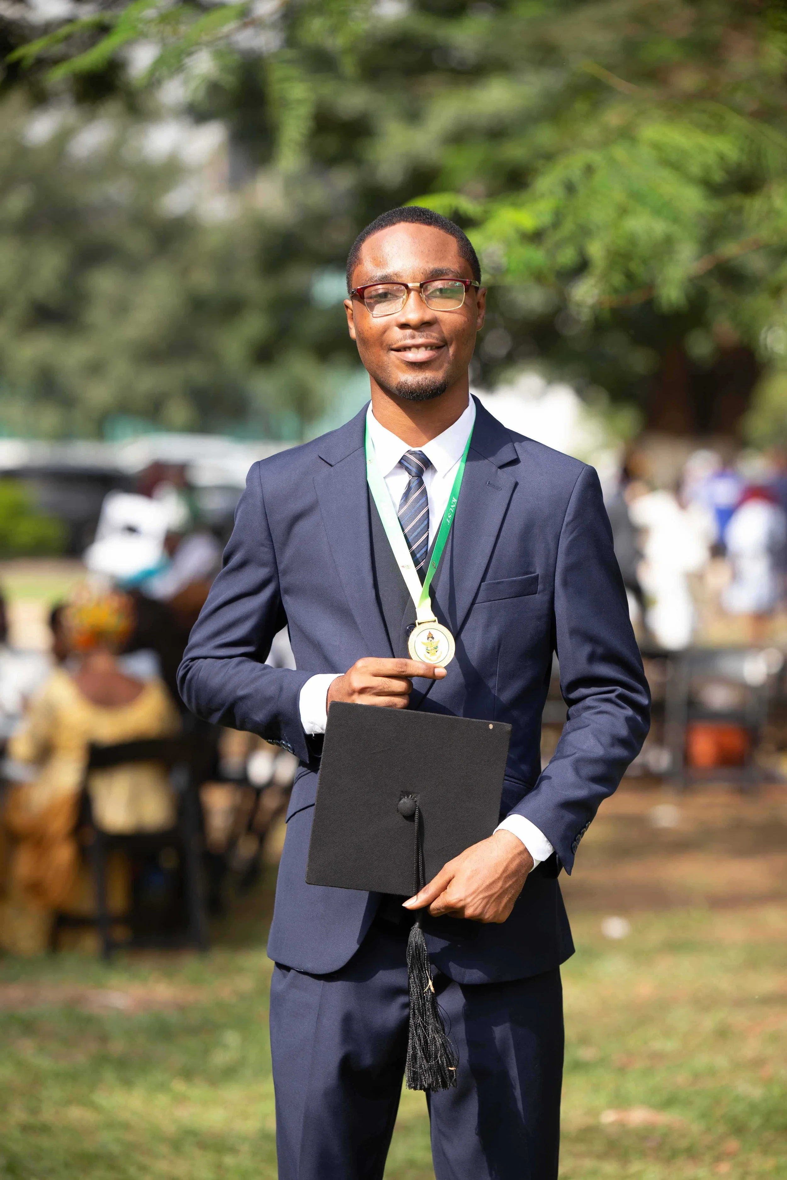 Man in a dark blue suit holding a framed graduation cap and wearing a medal around his neck at an outdoor graduation ceremony.