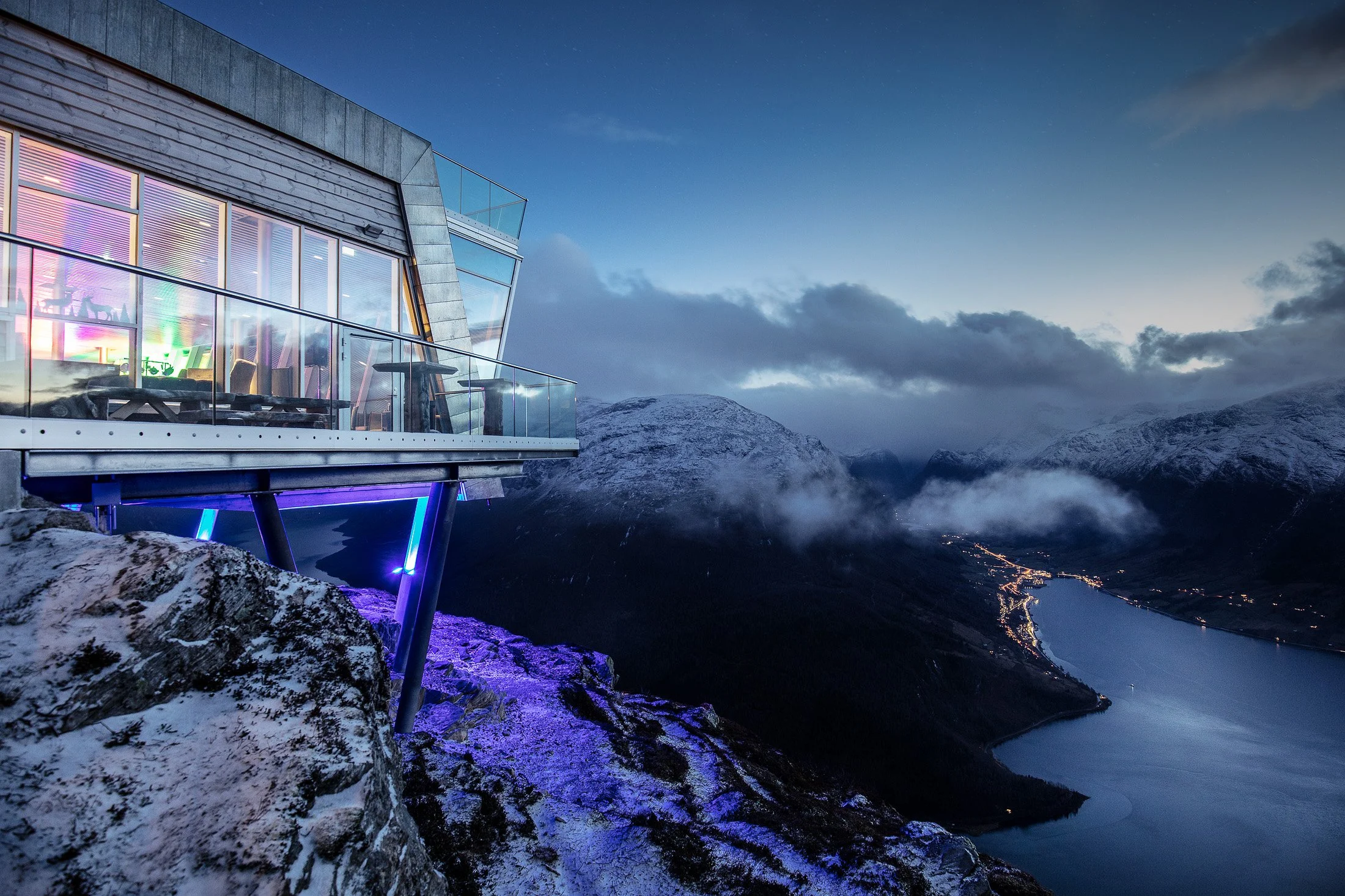 Modern glass building on a mountainside with a view of a fjord and snow-covered mountains at dusk.