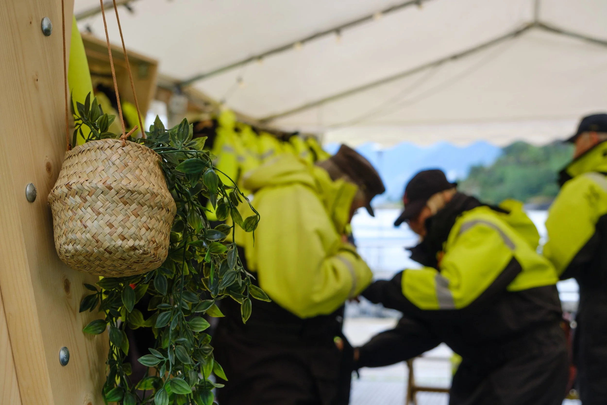 Close-up of a small woven basket with green leaves hanging on a wooden wall, with blurred people in yellow and black jackets in the background.