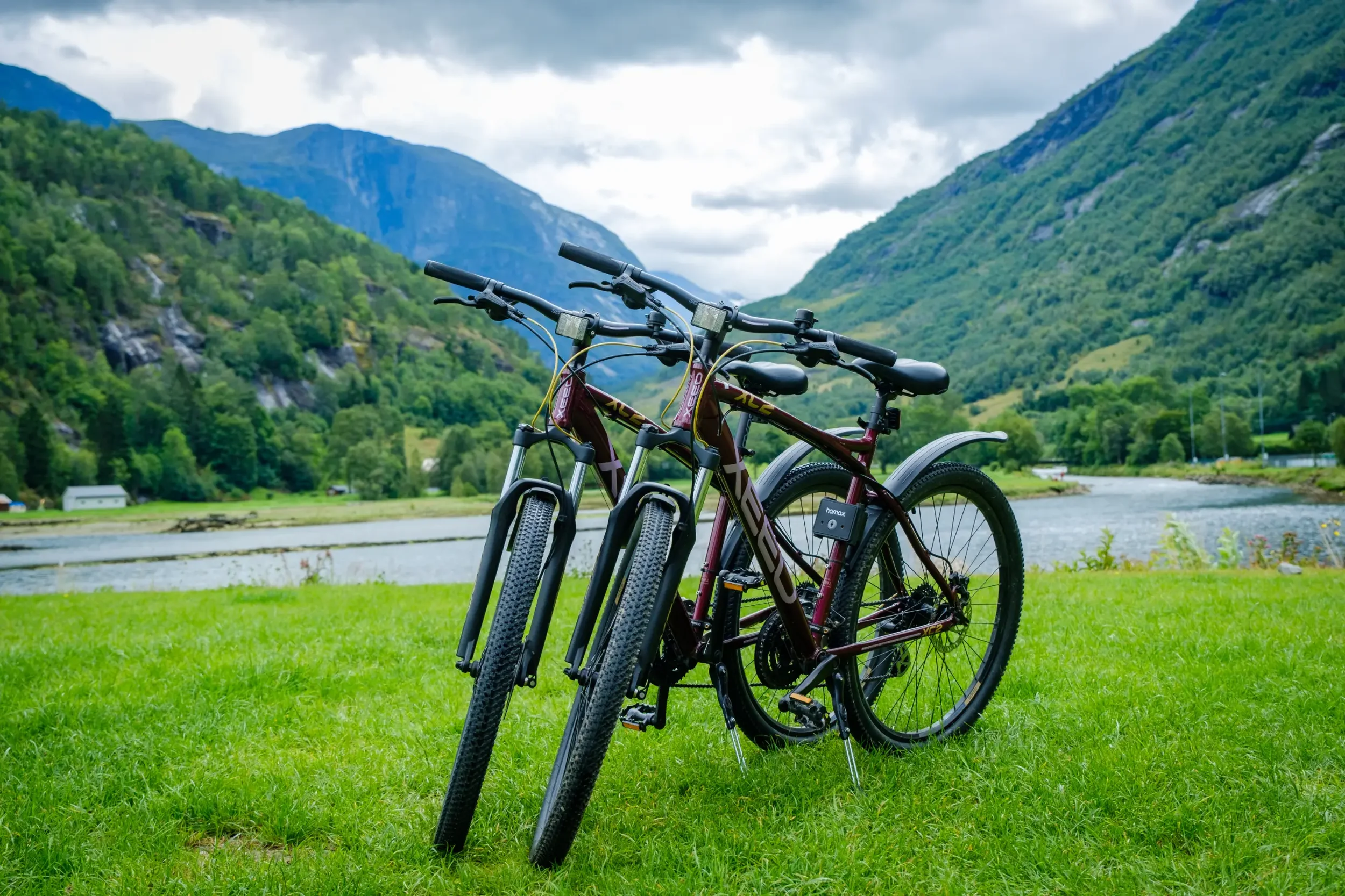 Two bikes parked on a grassy area near a river, with green mountains and cloudy sky in the background.
