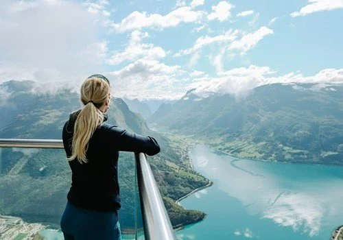 A woman with blonde hair in a ponytail stands on a balcony overlooking a scenic mountain and lake view.