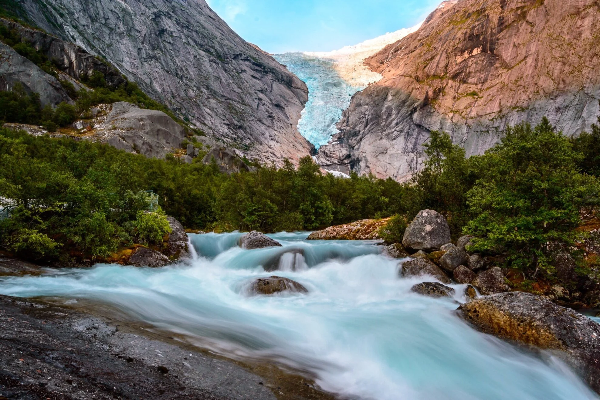 Flowing river with foam and rocks, green trees on shores, glacier at the mountain in the background, clear blue sky.