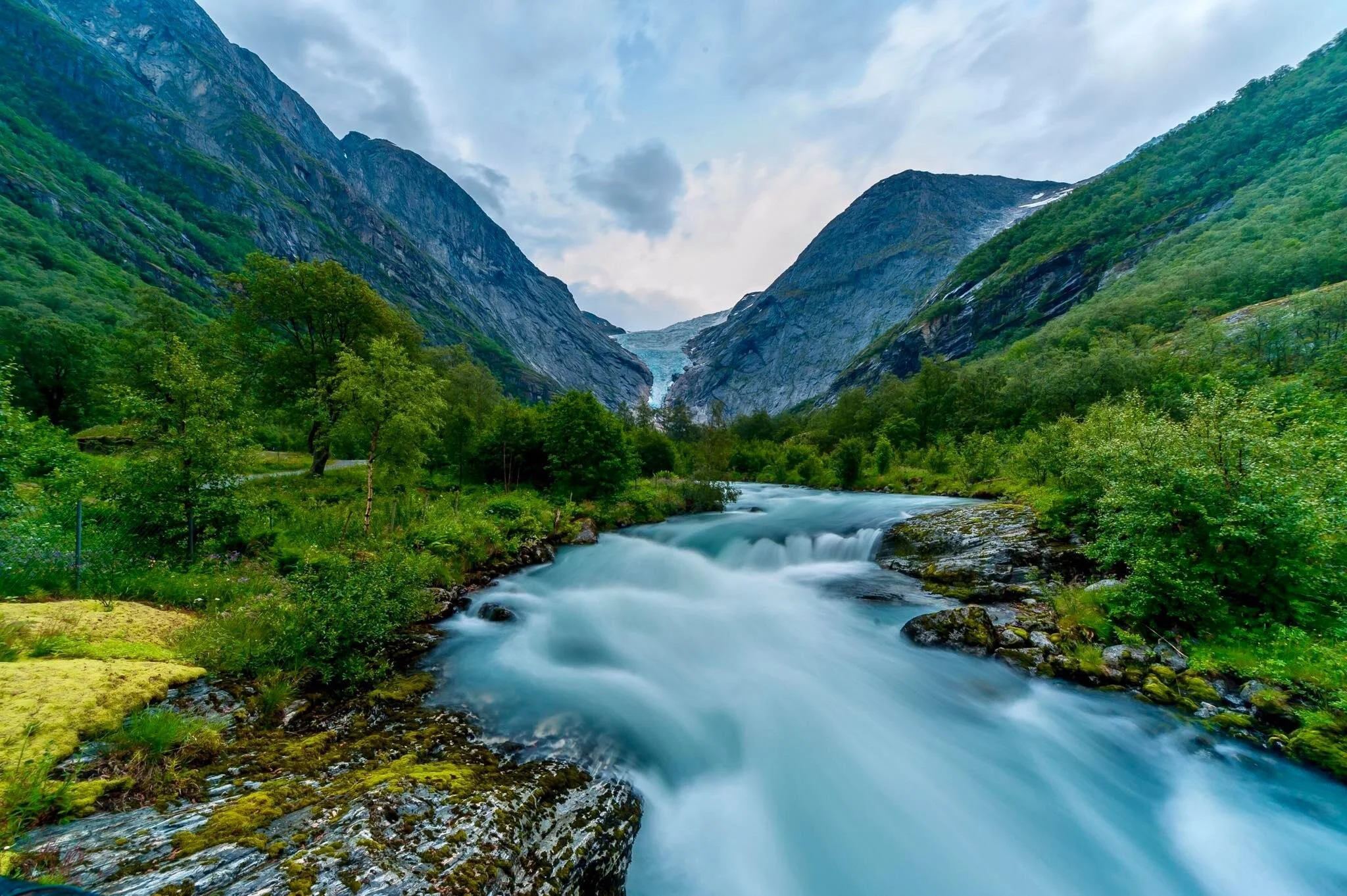 A scenic mountain landscape with a flowing river surrounded by lush green trees and rocky mountains, with a glacier visible in the background under a cloudy sky.