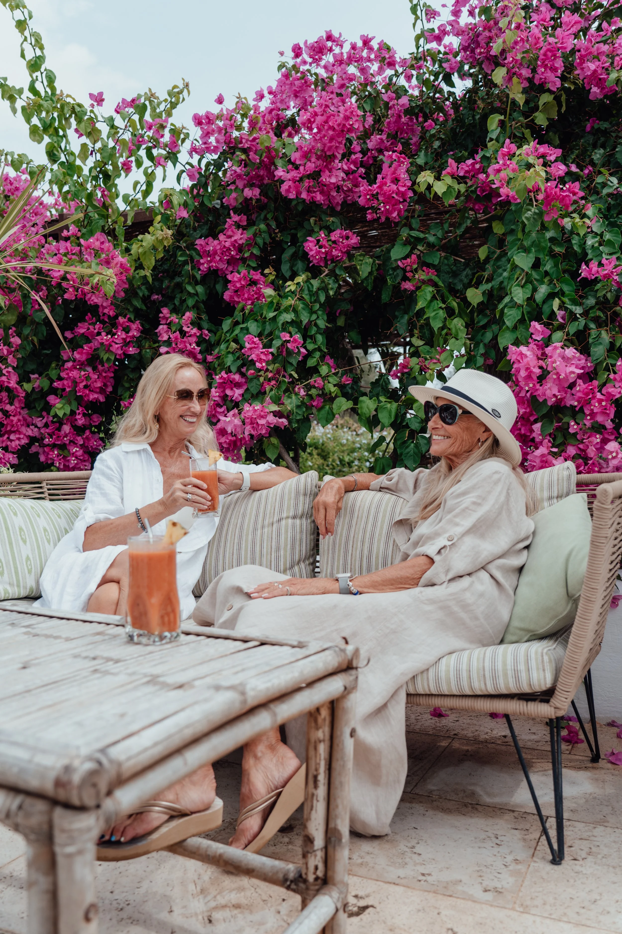 Two elderly women sitting outdoors on a couch, enjoying drinks, with pink bougainvillea flowers in the background, wearing summer clothing and sunglasses.