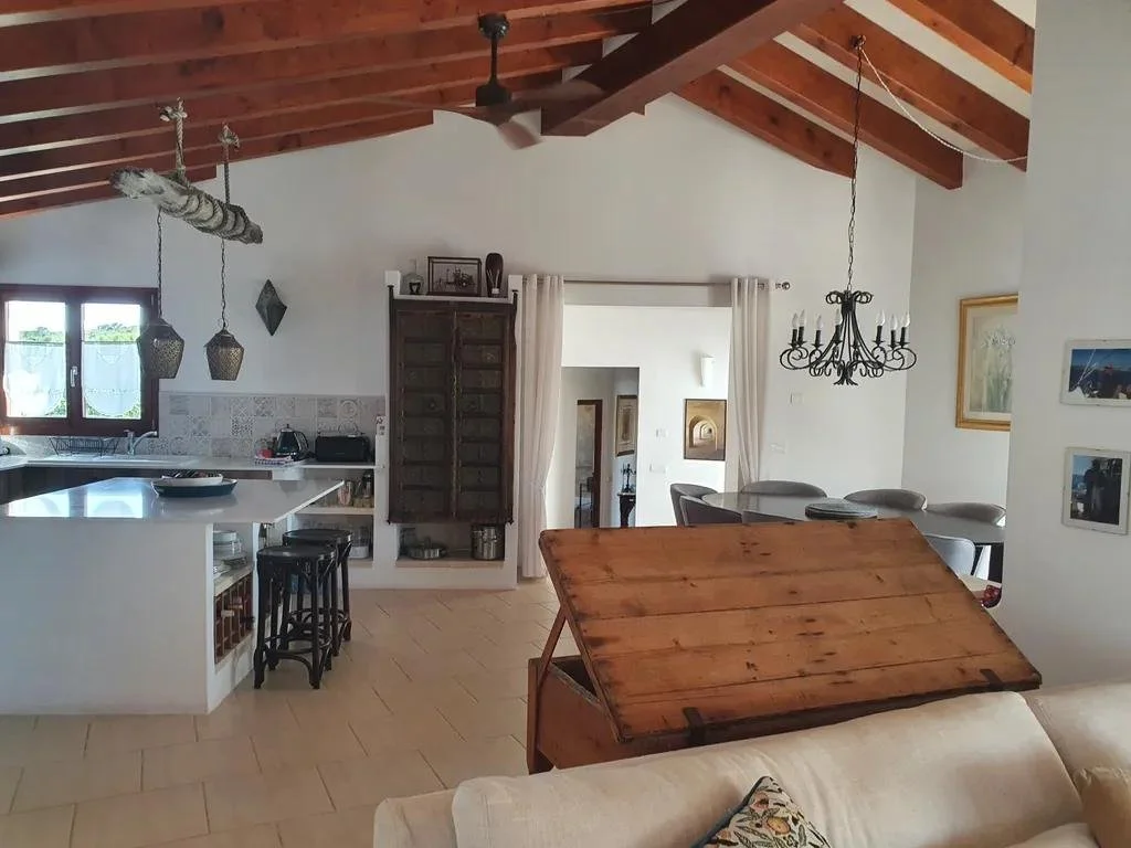Living room and kitchen with wooden ceiling beams, a dining table and chairs, a chandelier, a window with curtains, and a kitchen counter with stools.