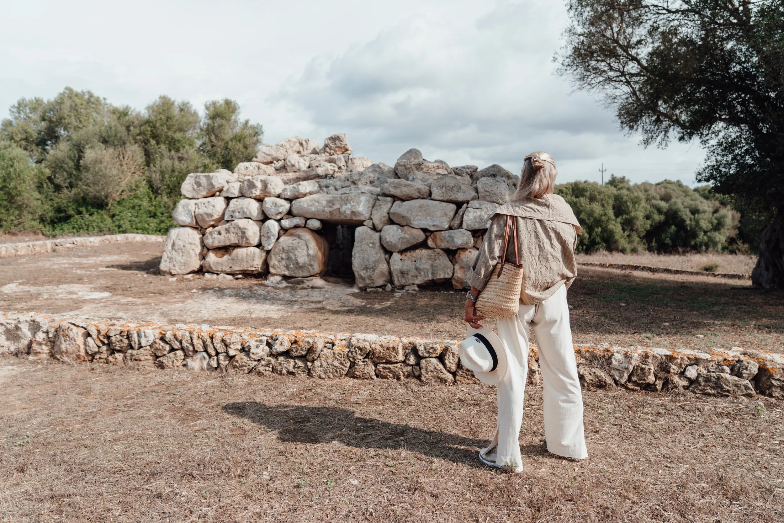 A woman with blonde hair wearing a beige jacket and white wide-leg pants, holding a straw bag and a white hat with a black band, stands near an ancient stone structure in a natural outdoor setting with trees and cloudy sky.