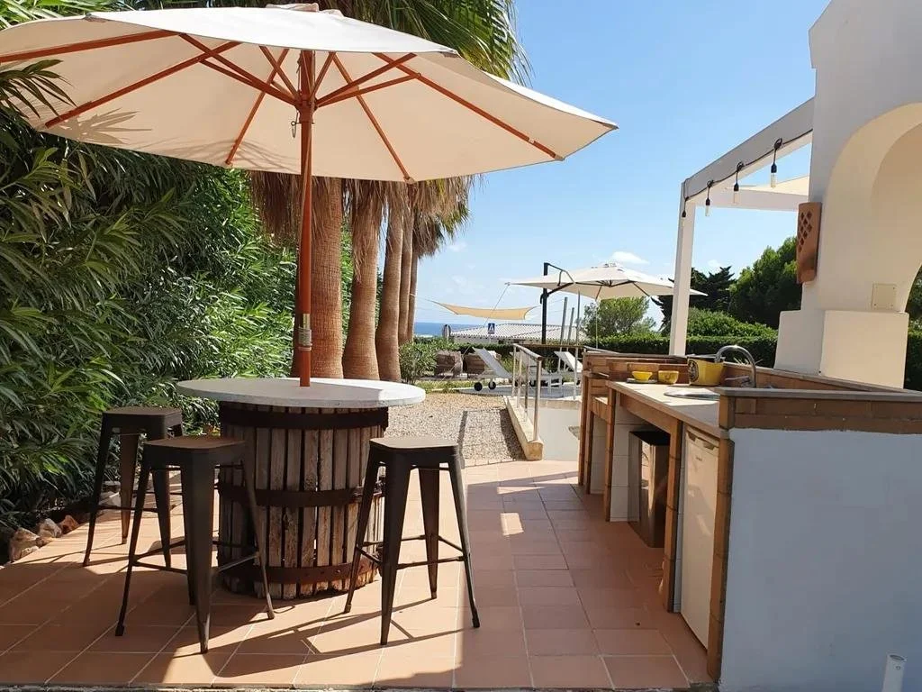 Outdoor kitchen and seating area with a round table, four bar stools, two large umbrellas, tropical greenery, and a view of the sea in the background.