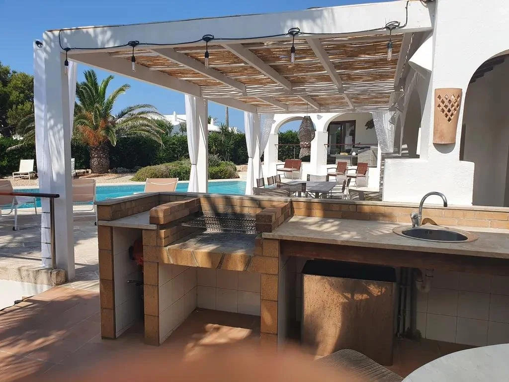 Outdoor pool area with a shaded patio, barbecue grill, sink, and seating. Palm trees and lounge chairs are visible in the background.