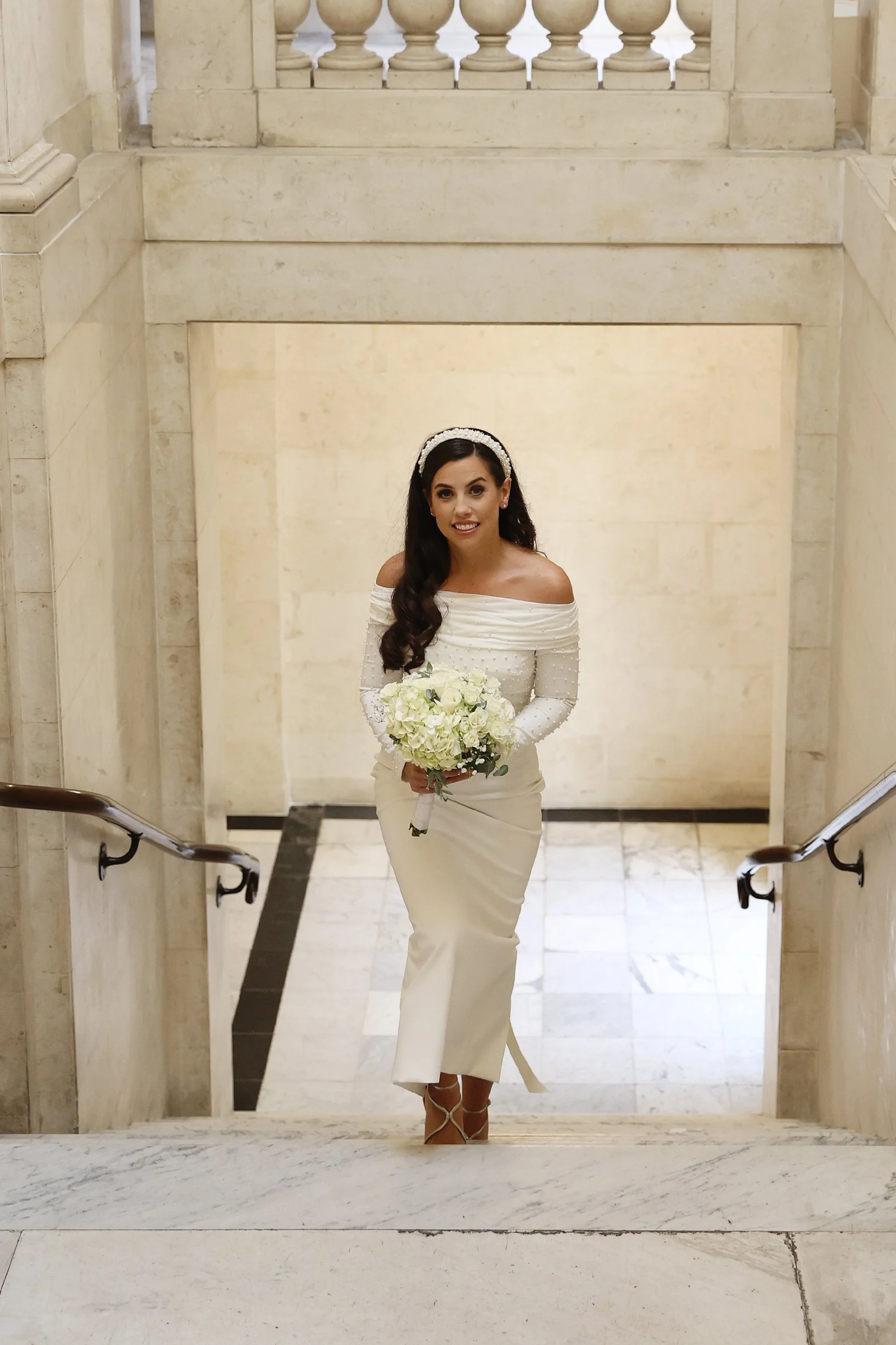 A woman in a strapless white wedding gown standing in a room surrounded by women, possibly helping her get dressed or ready.