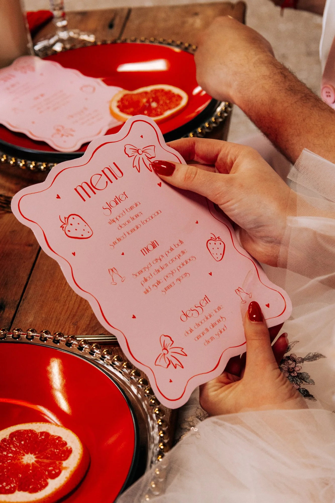 A woman holding a pink Valentine's Day themed menu card with red lettering and small hearts, strawberries, and ribbon illustrations, at a table with red and black plates and grapefruit slices.