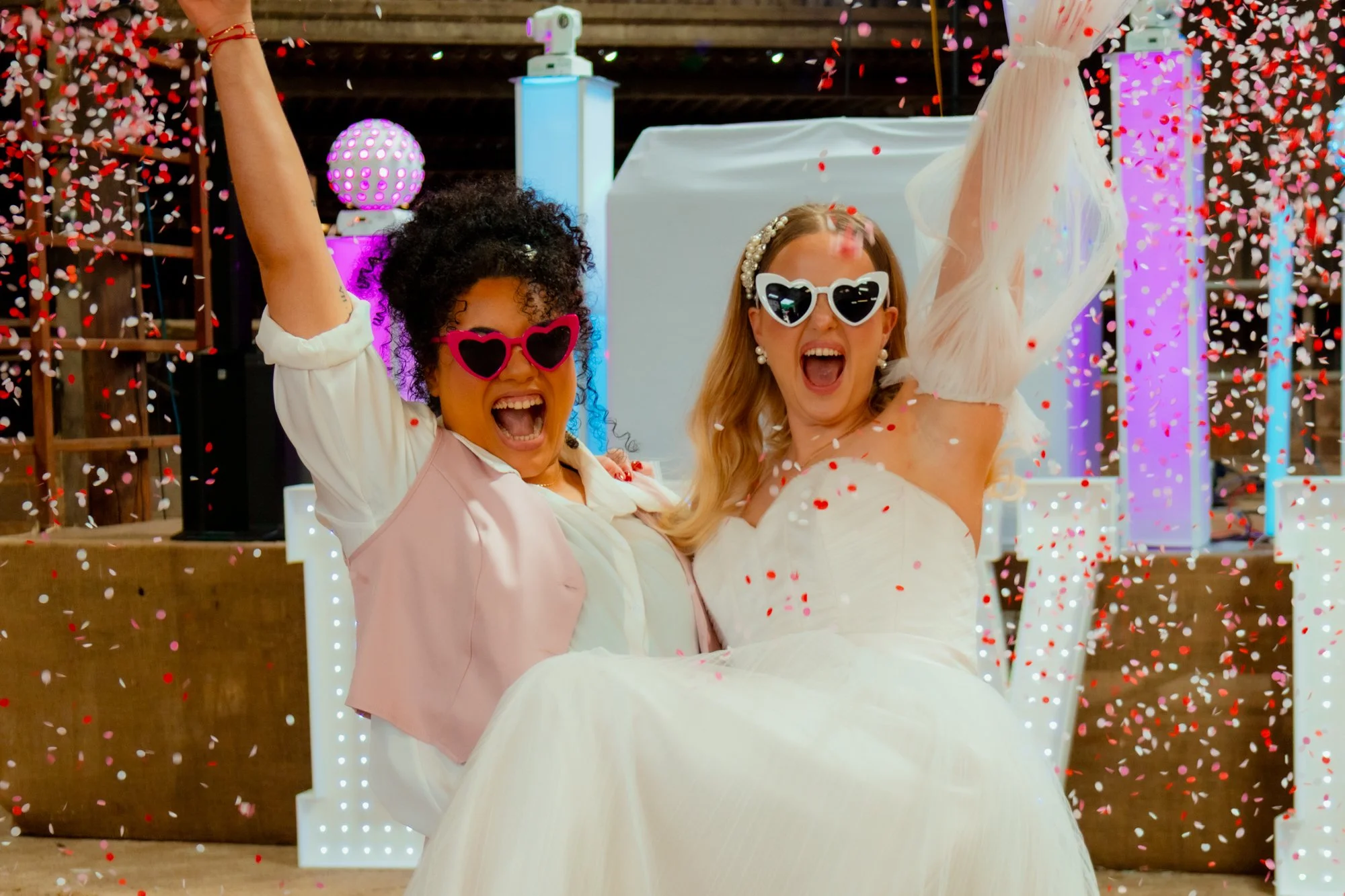 Two women, one in a wedding dress and the other in lighter colored clothes, celebrating with confetti and smiling at a wedding reception or party.