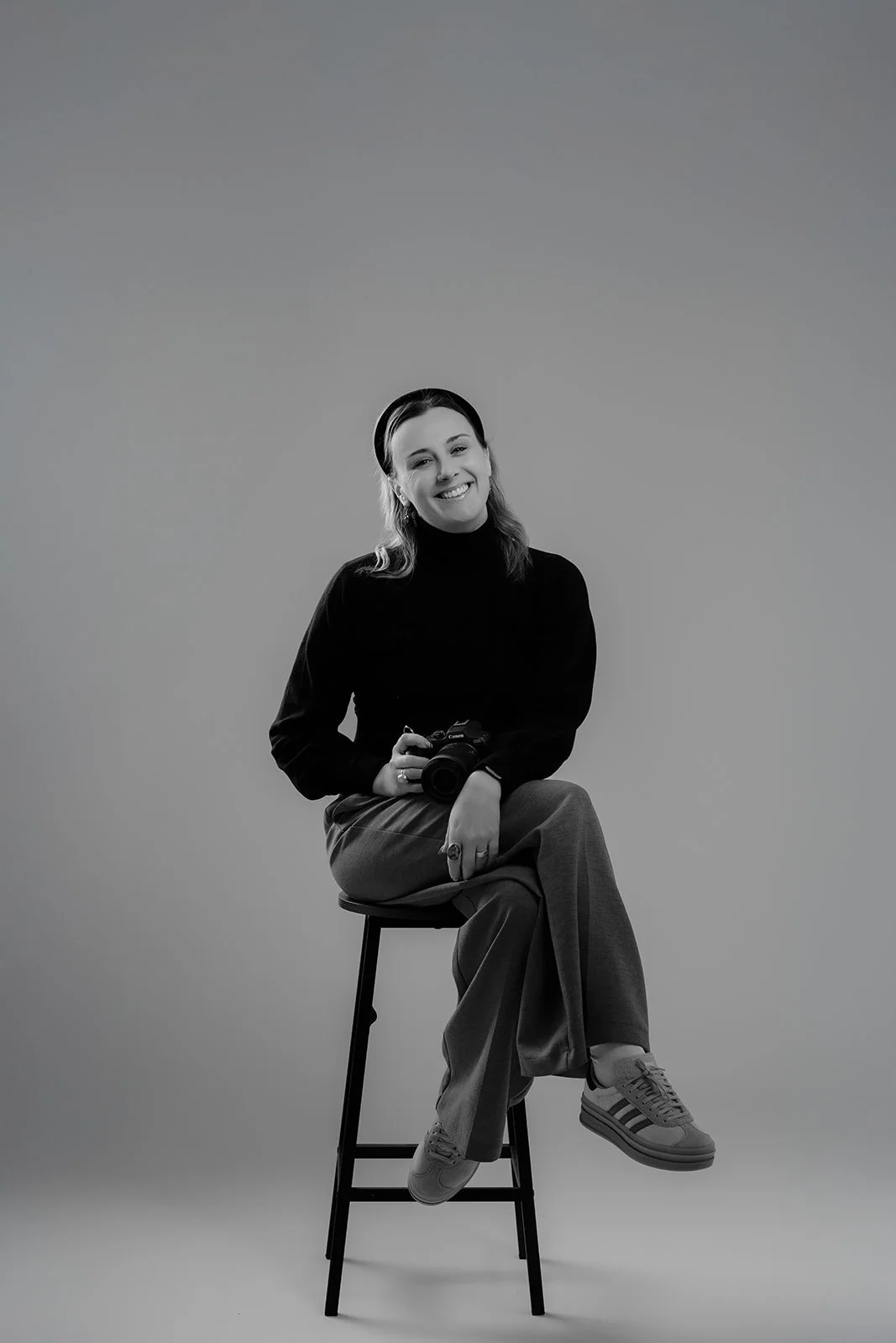 A young woman sitting on a stool, holding a camera, smiling, with her legs crossed, dressed in a black sweater, wide-legged pants, and sneakers, against a plain background.