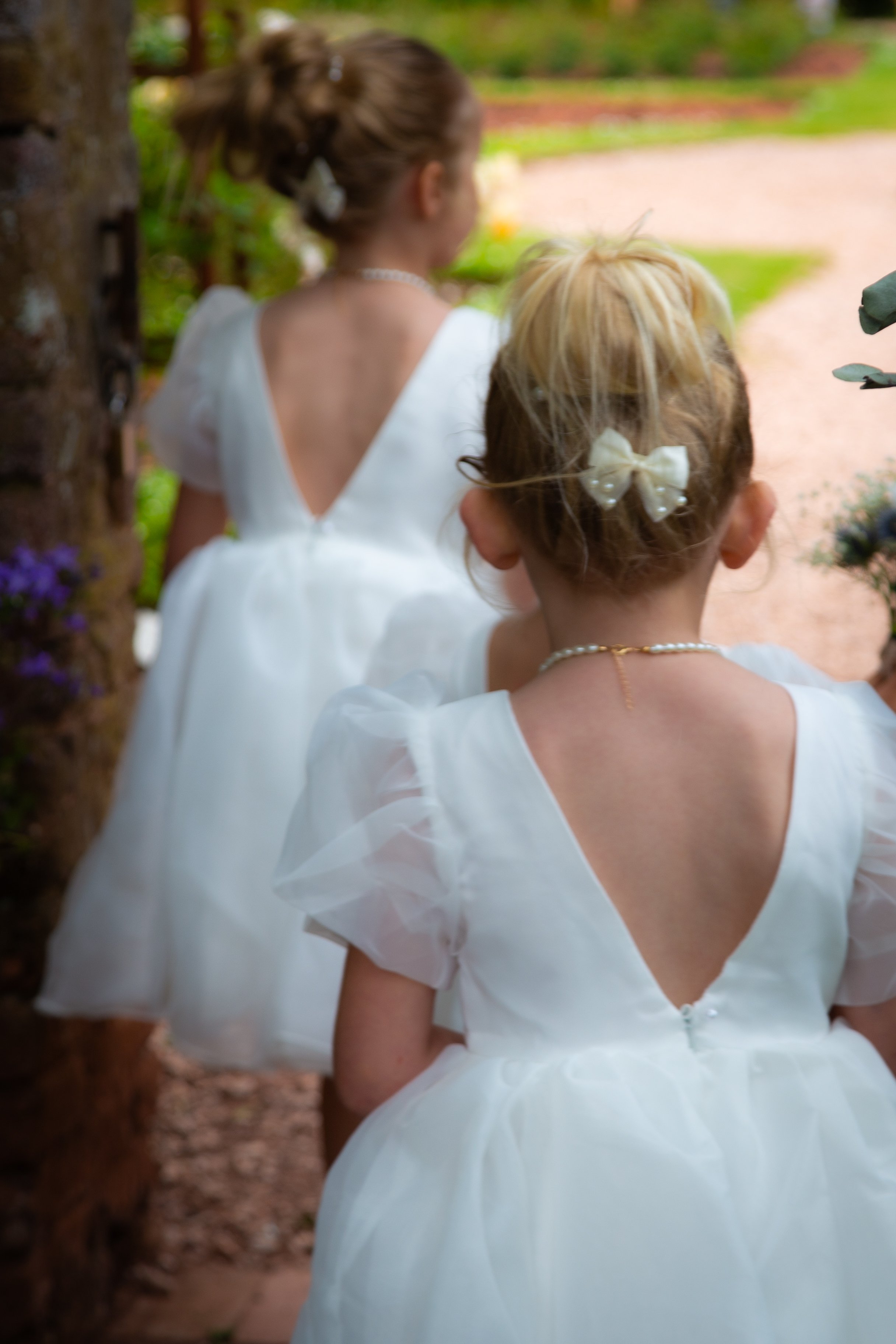 Two young girls dressed in white dresses with puffy sleeves, seen from behind, outdoors on a garden path. The girl in front has a bow in her hair, and the girl behind has a pearl necklace and a hair accessory, possibly for a special occasion or cerem