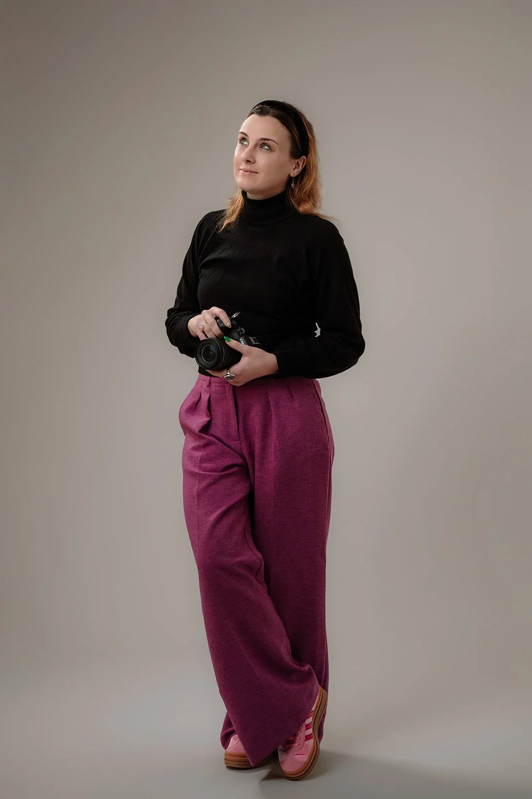 A woman standing against a neutral background holding a camera, wearing a black turtleneck, wide purple pants, and pink shoes.