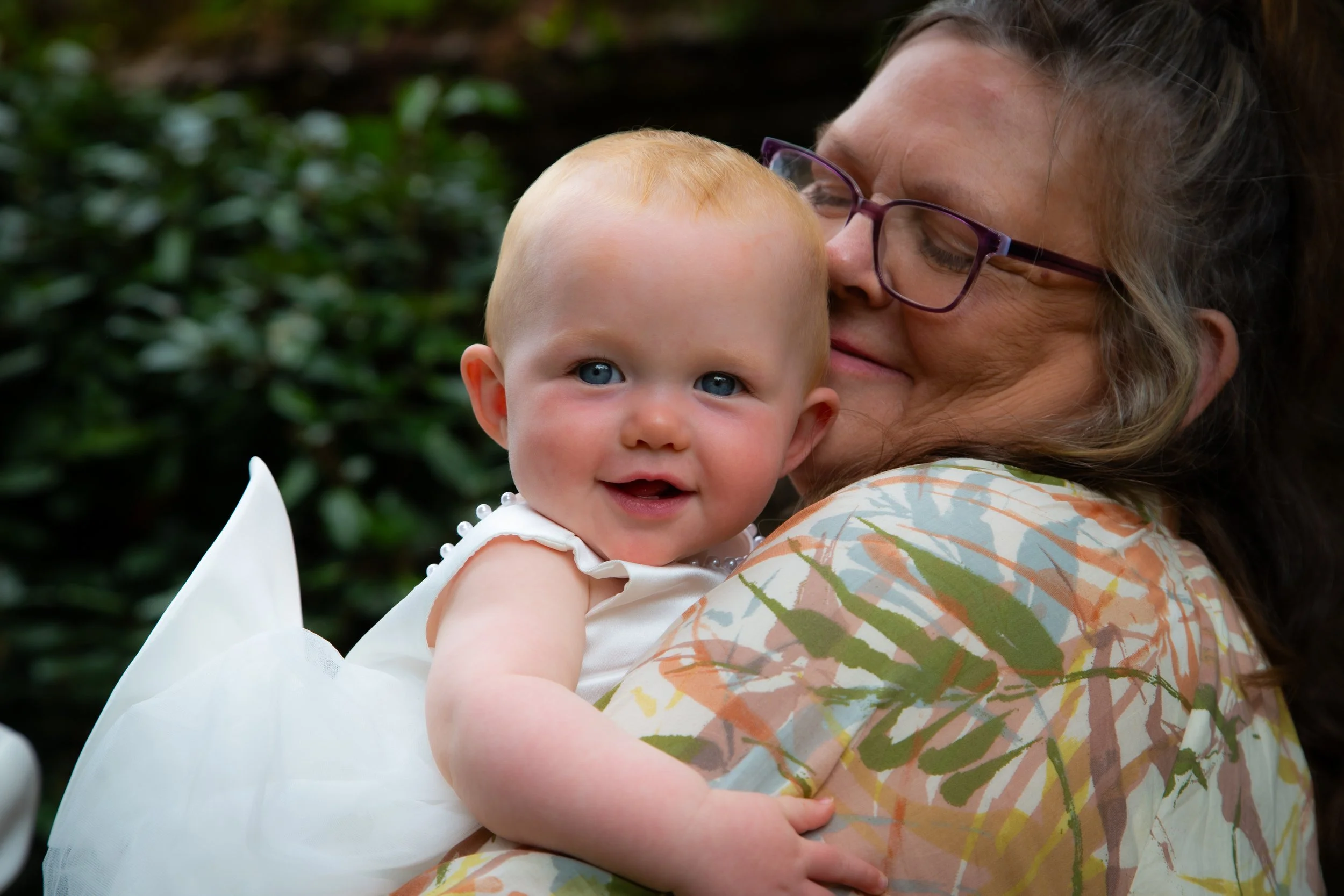 A woman with glasses hugging a smiling baby girl with blue eyes and blonde hair, outdoors with greenery in the background.