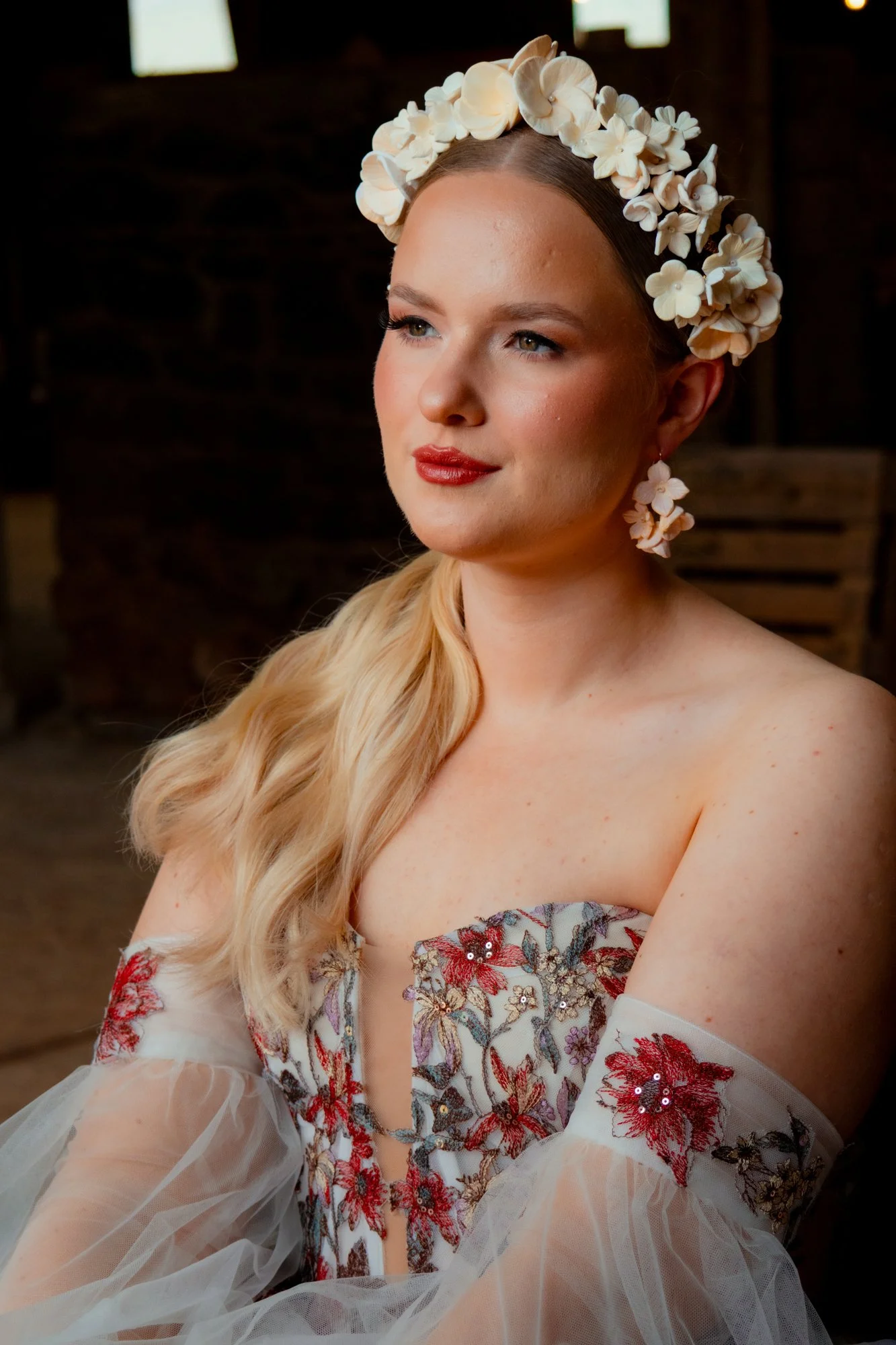 A woman with blonde hair in loose curls wearing floral earrings and a floral headband, dressed in a strapless embroidered dress with sheer sleeves.