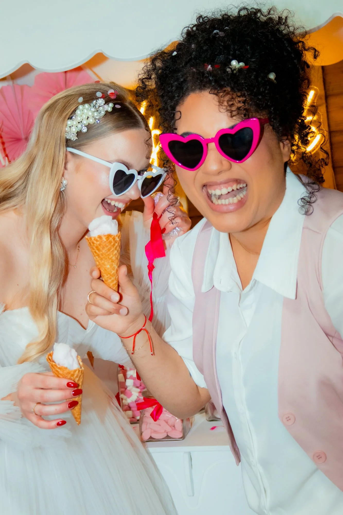 Two women wearing heart-shaped sunglasses, smiling and enjoying ice cream cones at a party.
