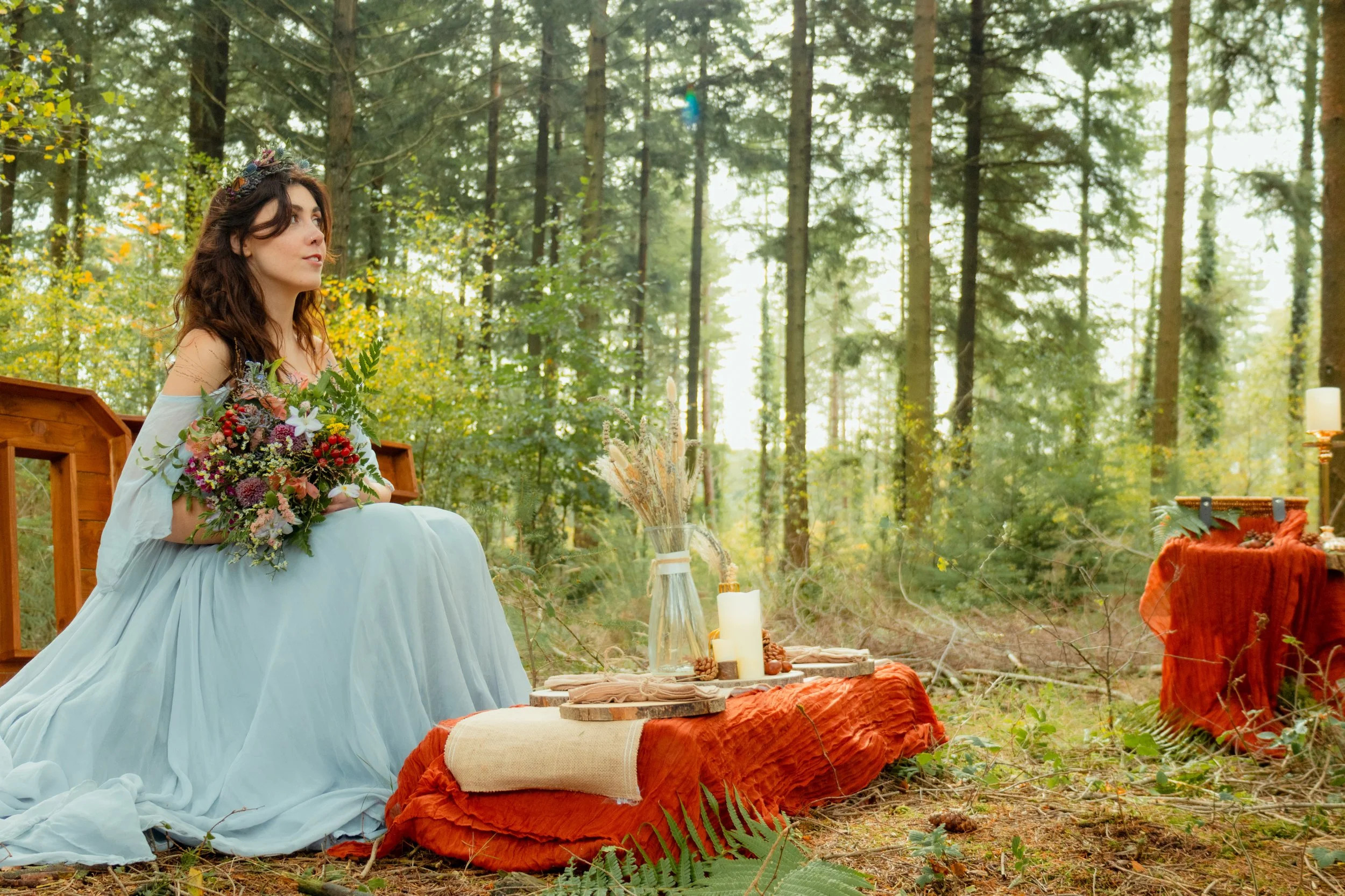 A woman in a flowing light blue dress sitting on the ground in a forest setting, holding a bouquet of flowers, with decorative tables, candles, and foliage around her.