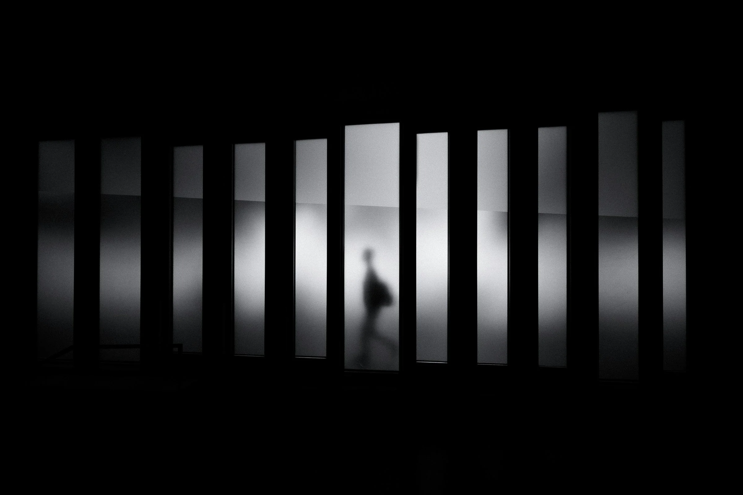 blurred silhouette of a person walking, seen through vertical window blinds or bars in a dark room.