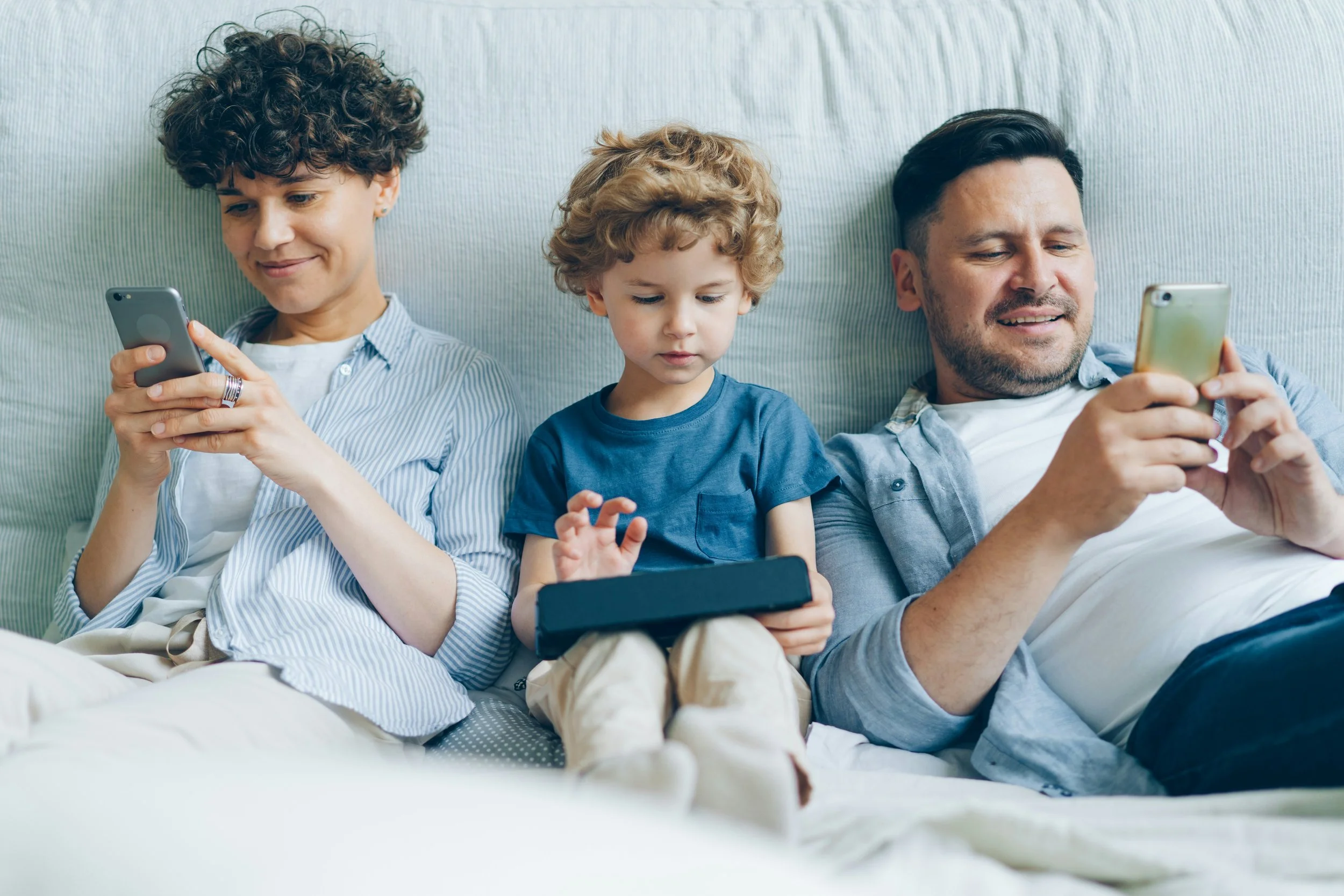 A mom and dad are sitting on a bed with their son, they are all looking at their devices