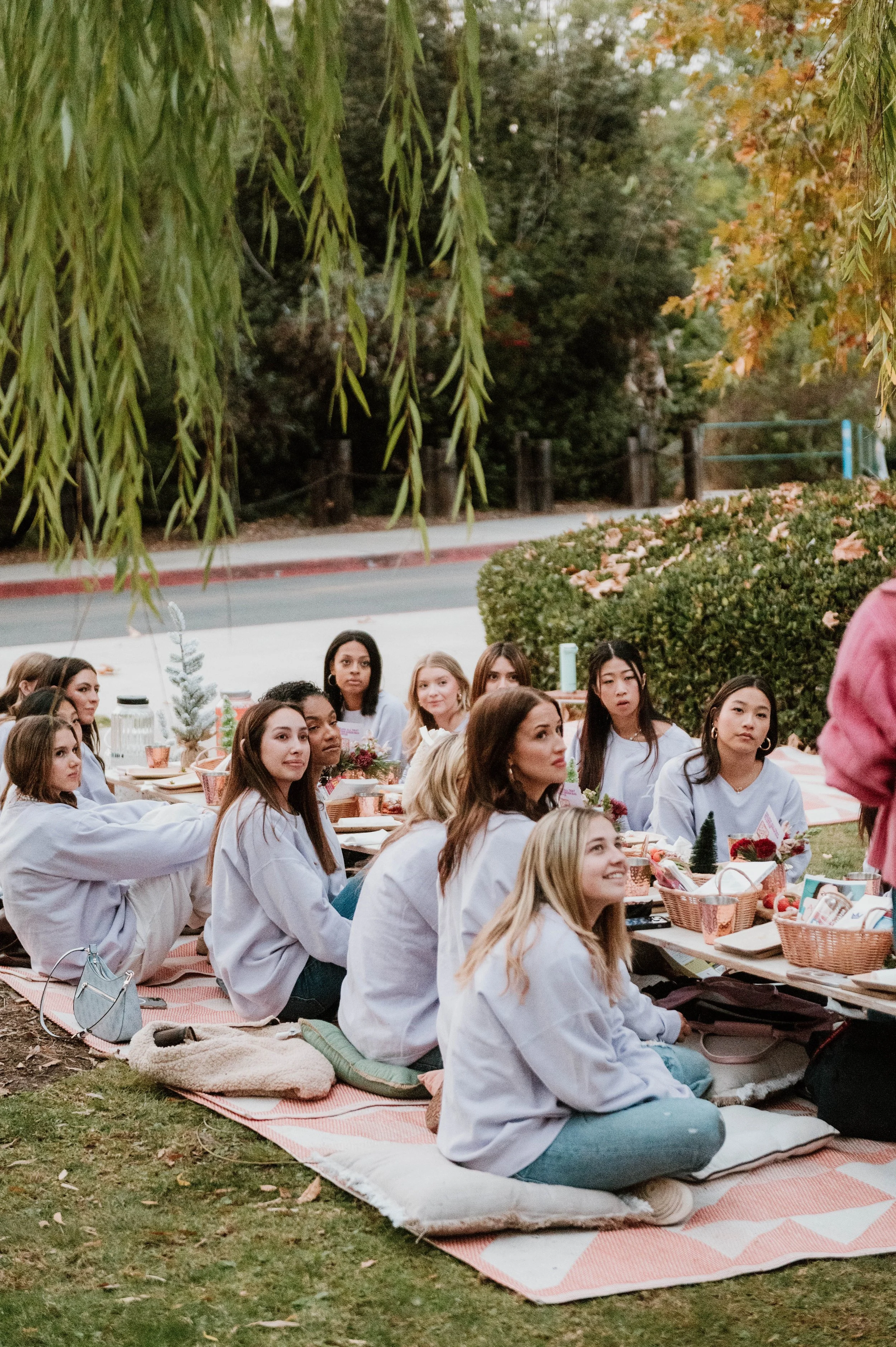 A group of people sitting on a picnic blanket outdoors, listening to someone in a pink jacket. They are surrounded by trees and bushes, with some people taking notes or holding books.