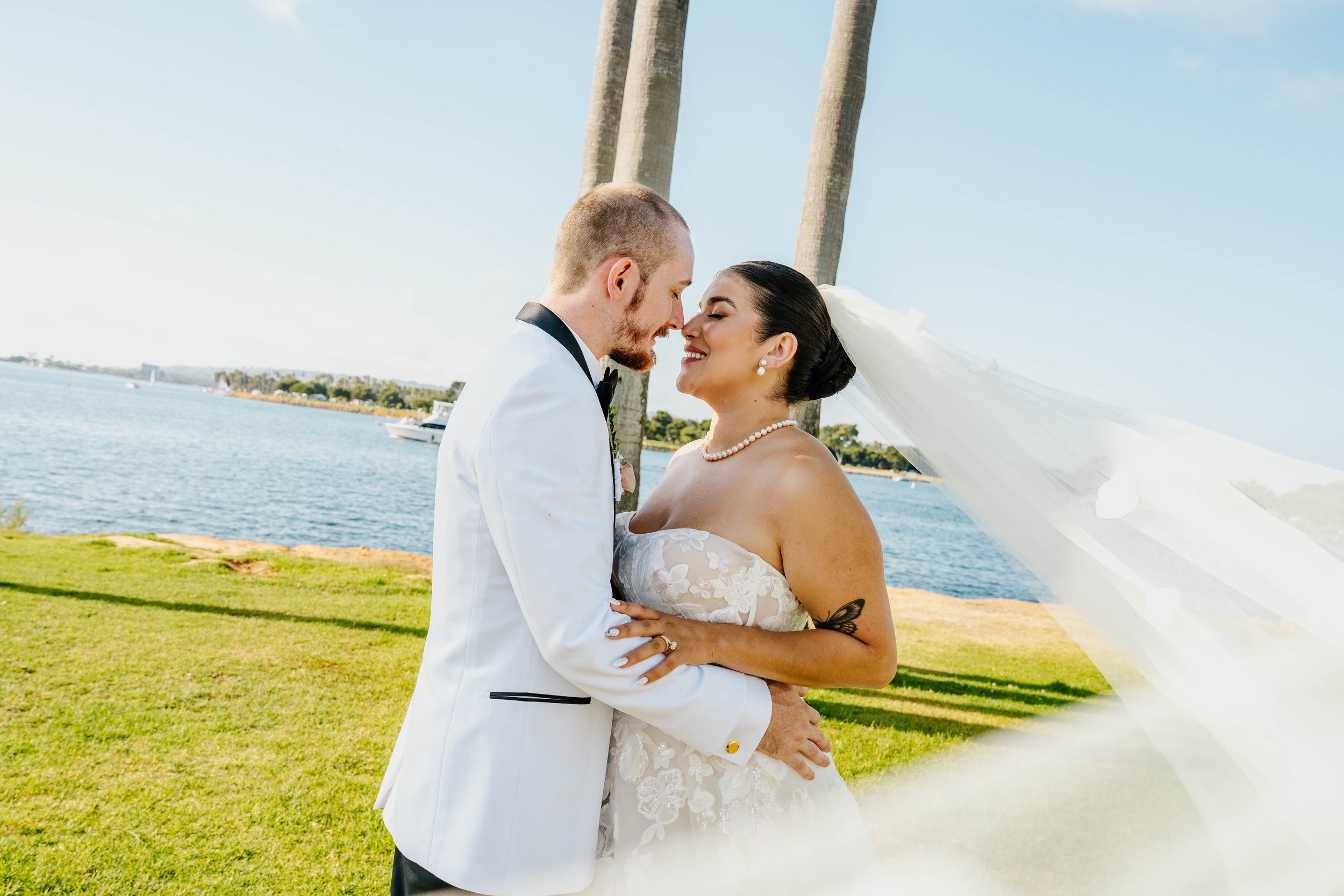 A newlywed couple, a man in a white tuxedo and a woman in a white lace wedding dress with a pearl necklace and earrings, share a tender moment with their foreheads touching and eyes closed, outdoors near water with a grassy area and palm trees in the background.