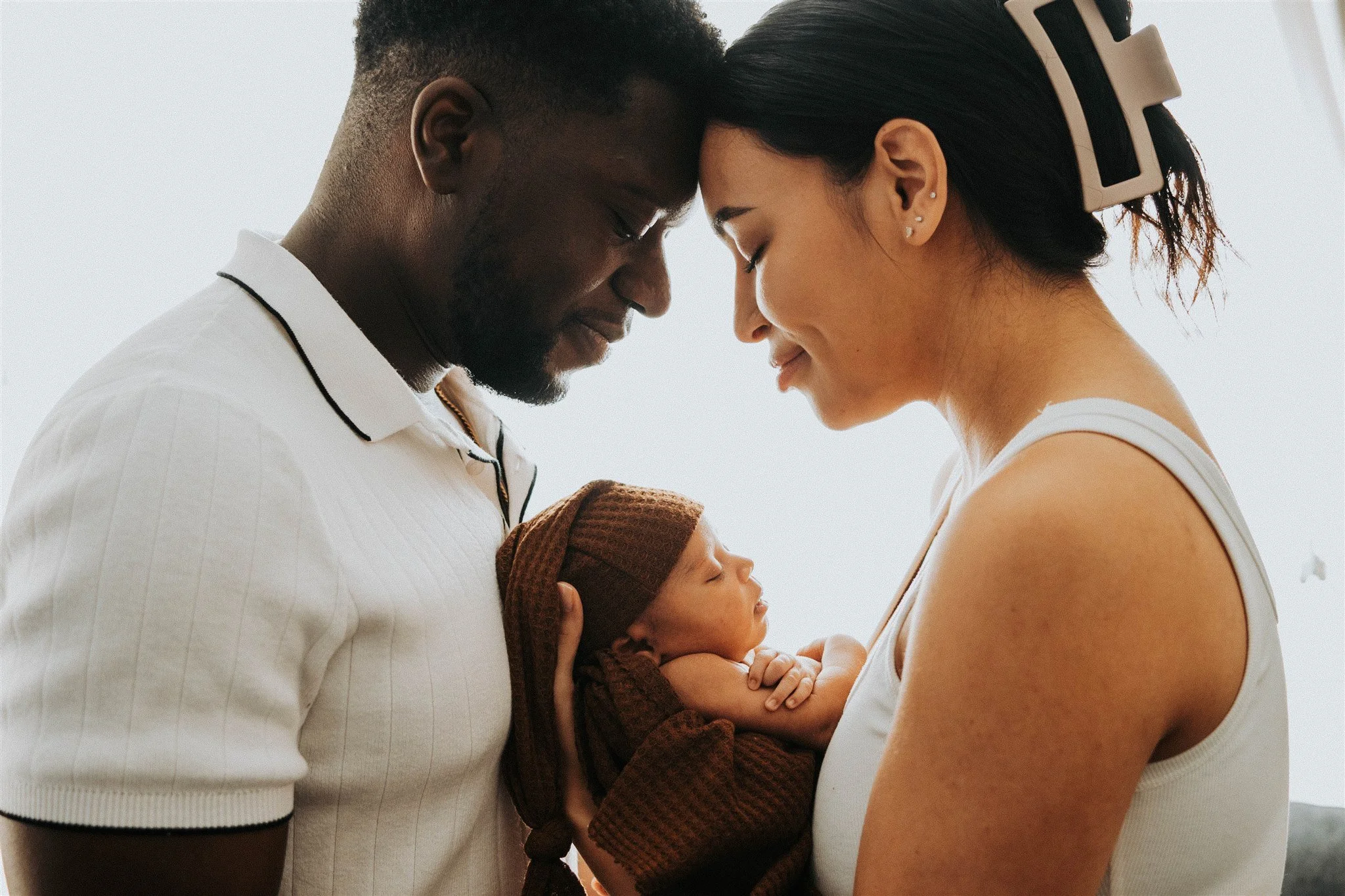 A family with a man, woman, and a baby, all with their foreheads touching and eyes closed, sharing a tender moment.