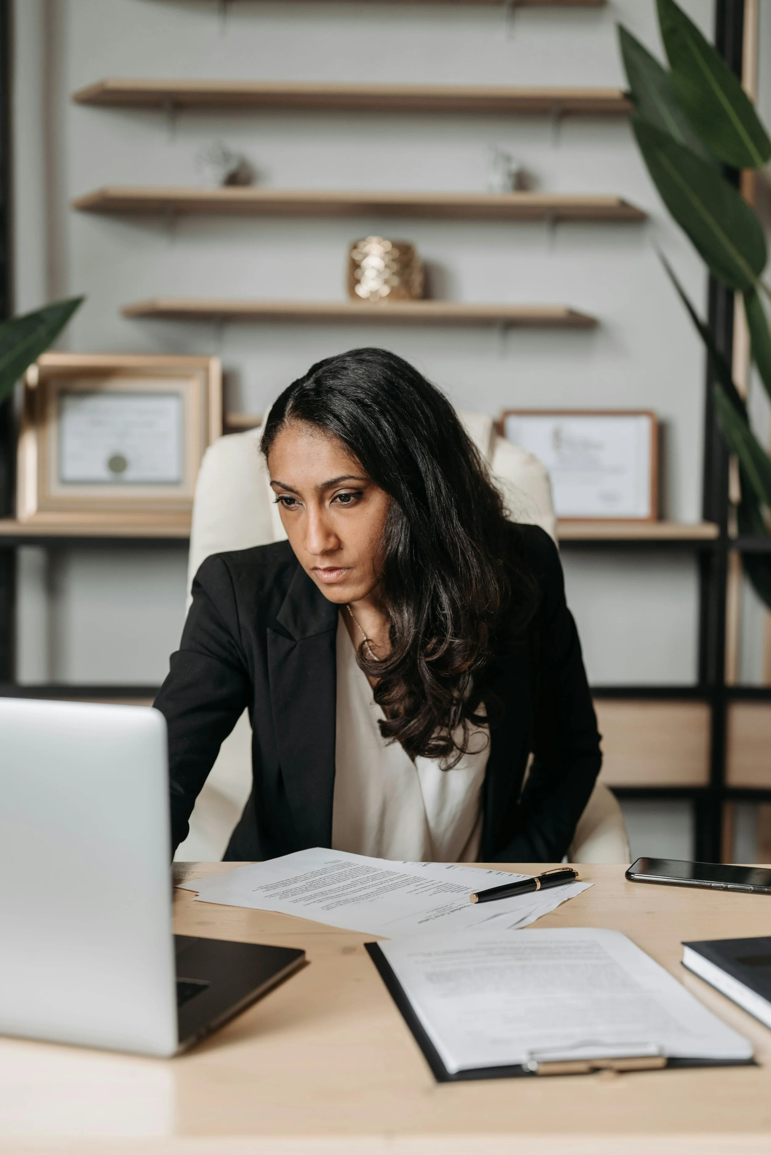 A woman in business attire sitting at a desk with documents, a pen, and a laptop, looking focused at her computer screen in an office setting with framed certificates and shelves in the background.