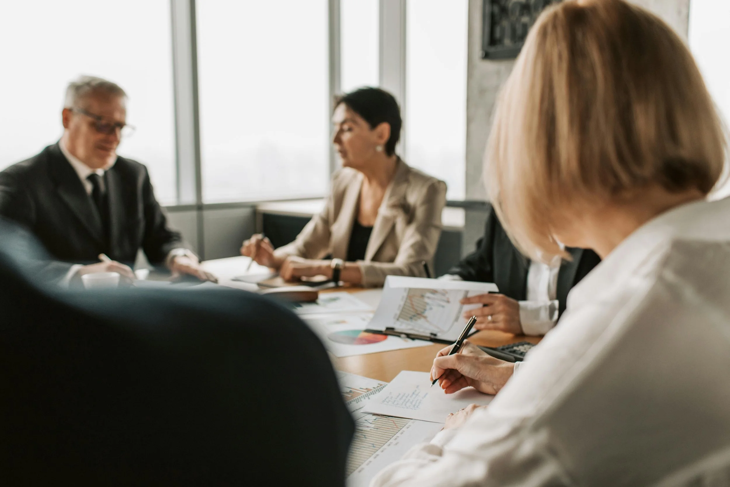 Business meeting with four professionals at a conference table, discussing documents and charts, in a bright office with large windows.