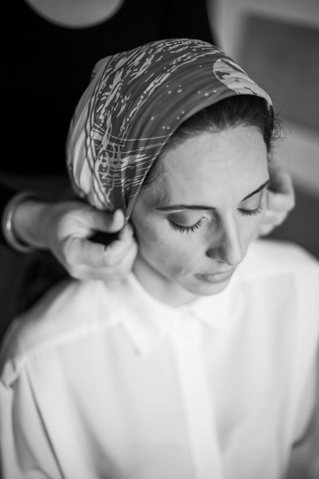 Une femme avec un bandana coloré sur la tête, dont la personne qui la coiffe ajuste le tissu, vue de profil avec les yeux fermés.