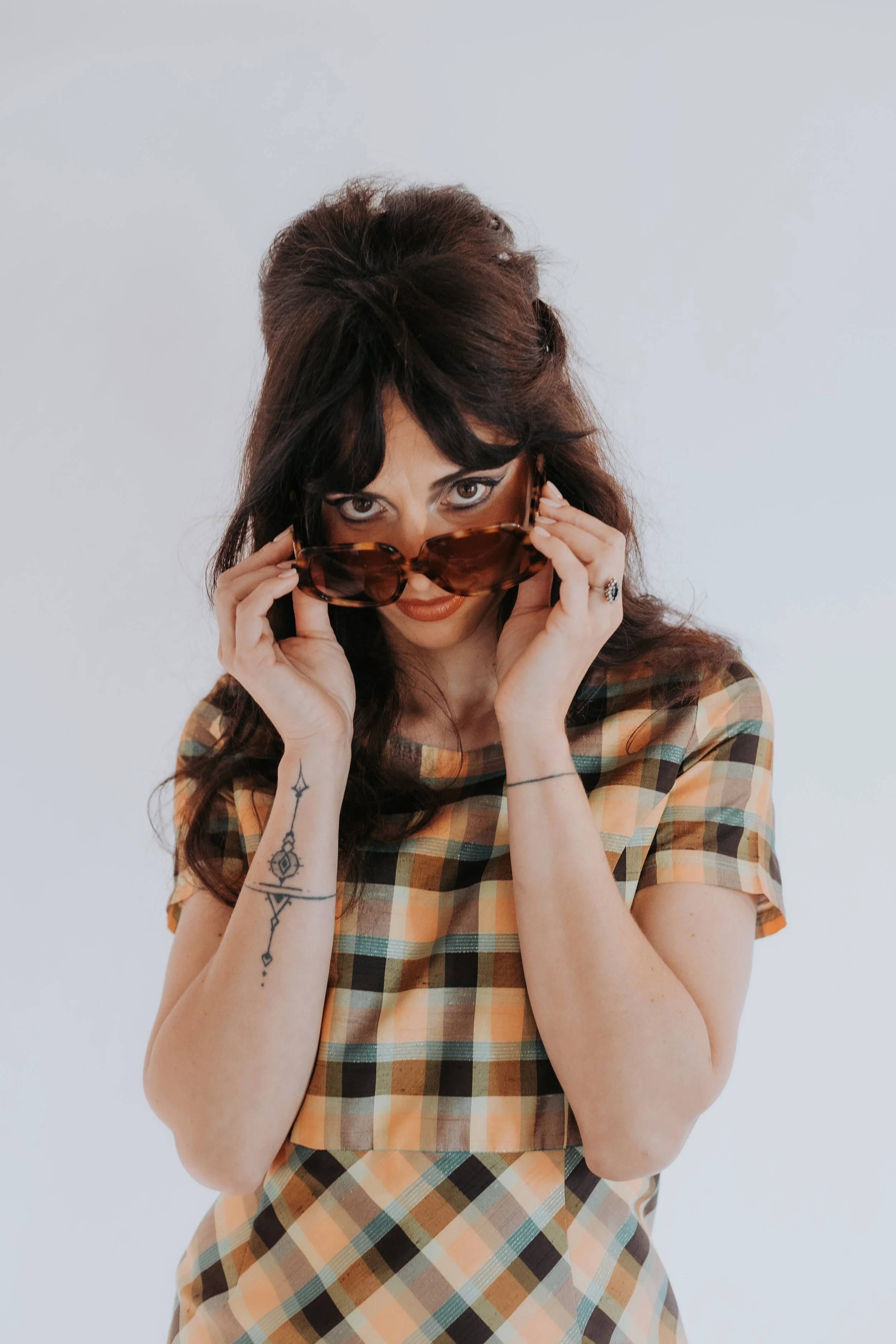 Jeune femme avec un tatouage sur le bras, portant des lunnettes de soleil, en vêtement à carreaux or jaune, pose devant un fond blanc.