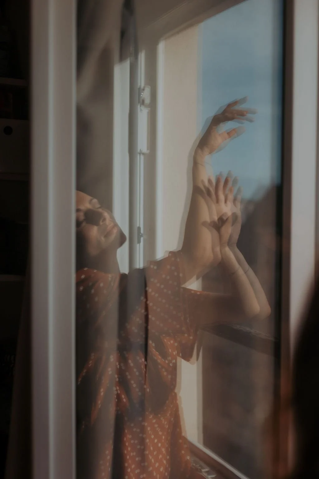 Une femme vêtue d'une robe à pois regarde par une fenêtre, sa réflexion est visible sur la vitre.