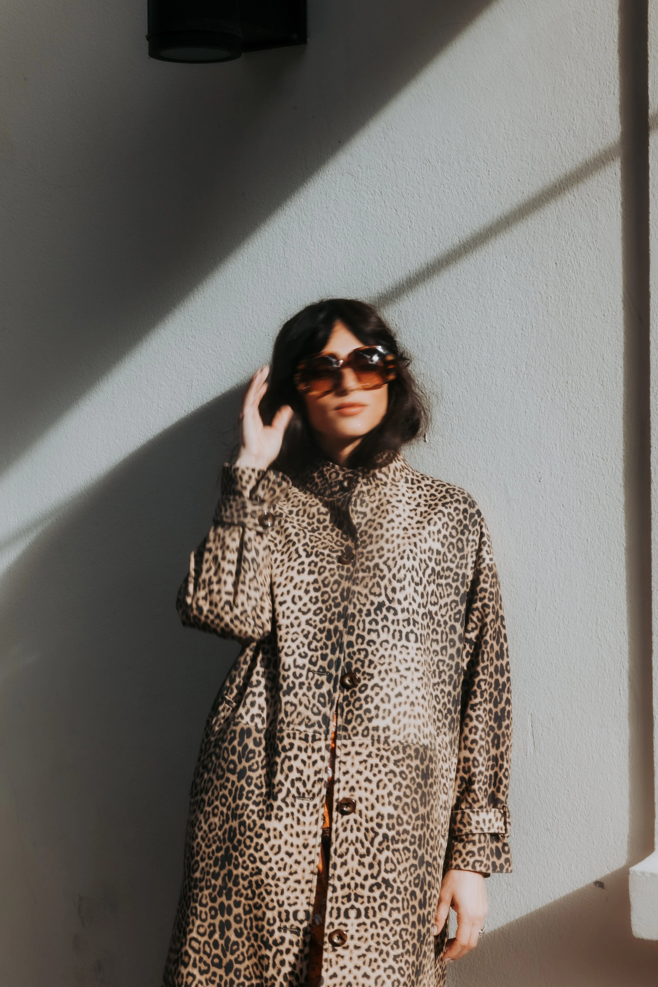 Femme portant une robe à imprimé léopard, portant des lunettes de soleil, se tenant devant un mur avec une lumière du soleil créant une ombre.