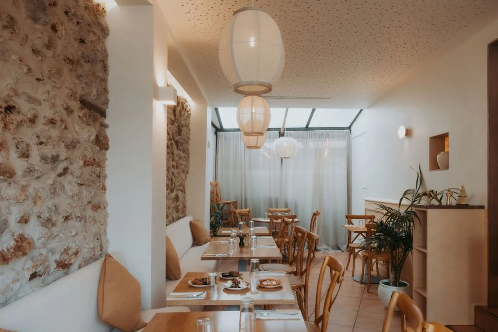 Intérieur d'une salle à manger lumineuse avec murs en pierre, tables en bois, chaises en bois, lampes suspendues en papier, plantes vertes et rideaux blancs