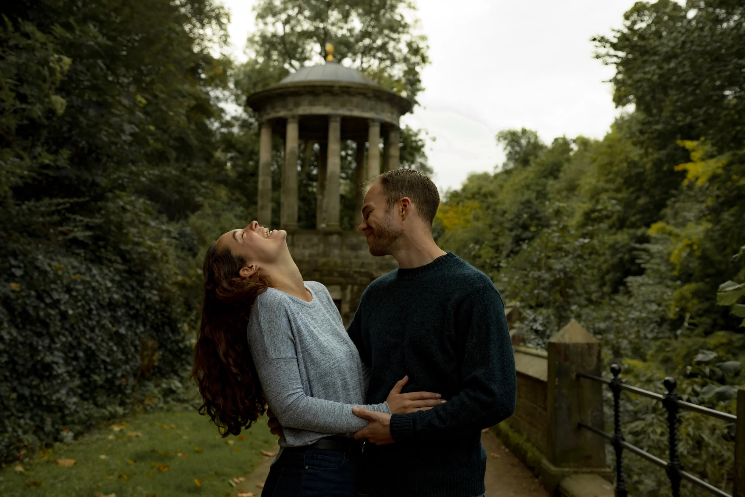 A couple hugging and laughing in a park with a gazebo in the background.