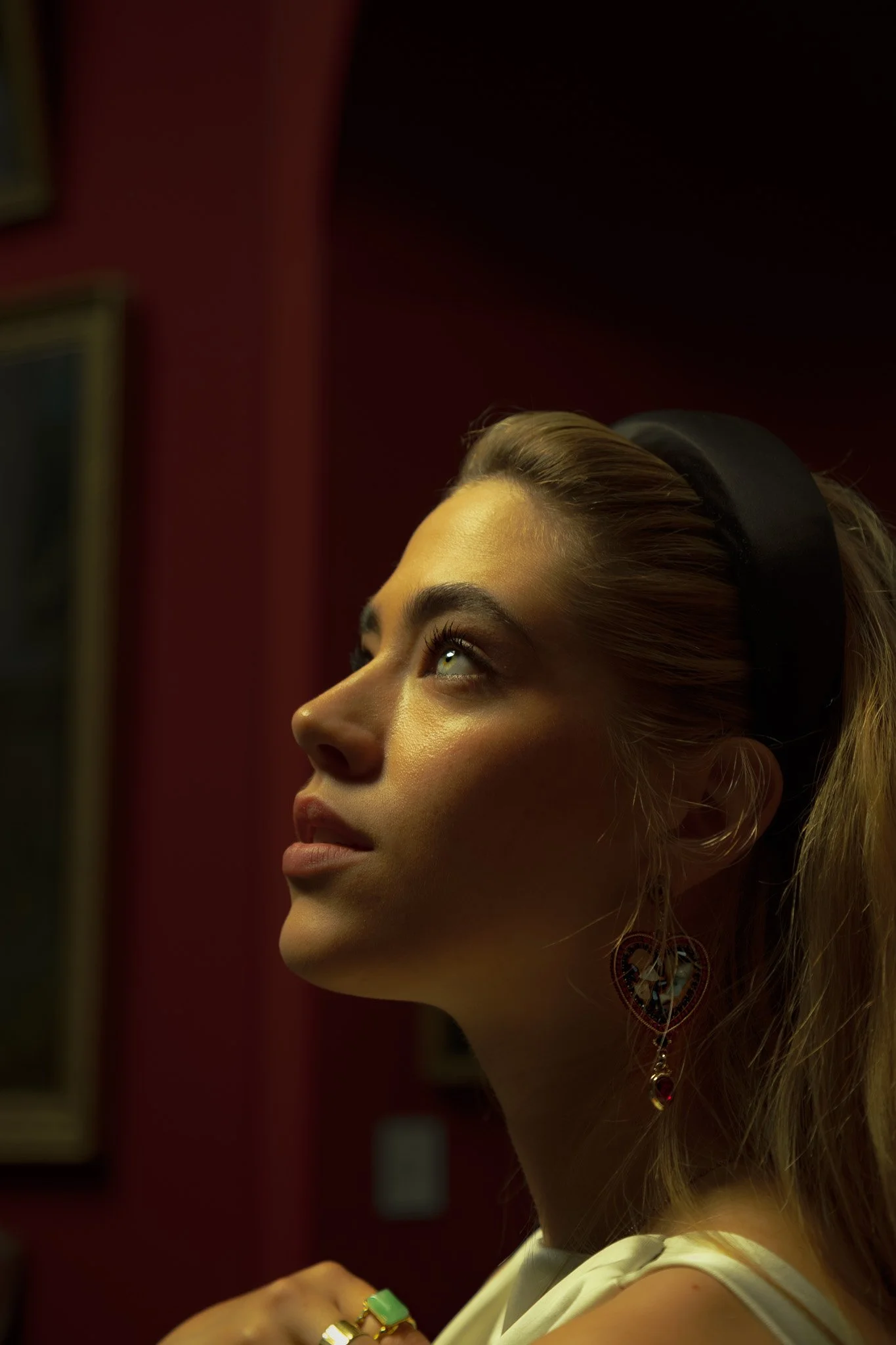 A young woman with blonde hair and creative makeup looking upward, wearing colorful earrings, a headband, and a ring, with a red wall and framed pictures in the background.